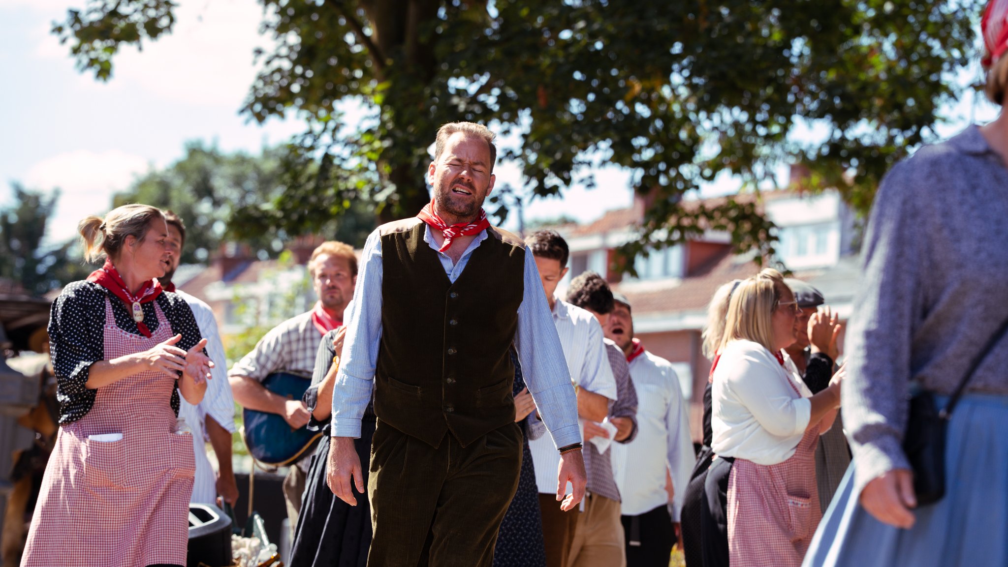 Group of people, dressed in traditional or vintage clothing, participating in an outdoor event under a large tree, with some making expressive gestures.