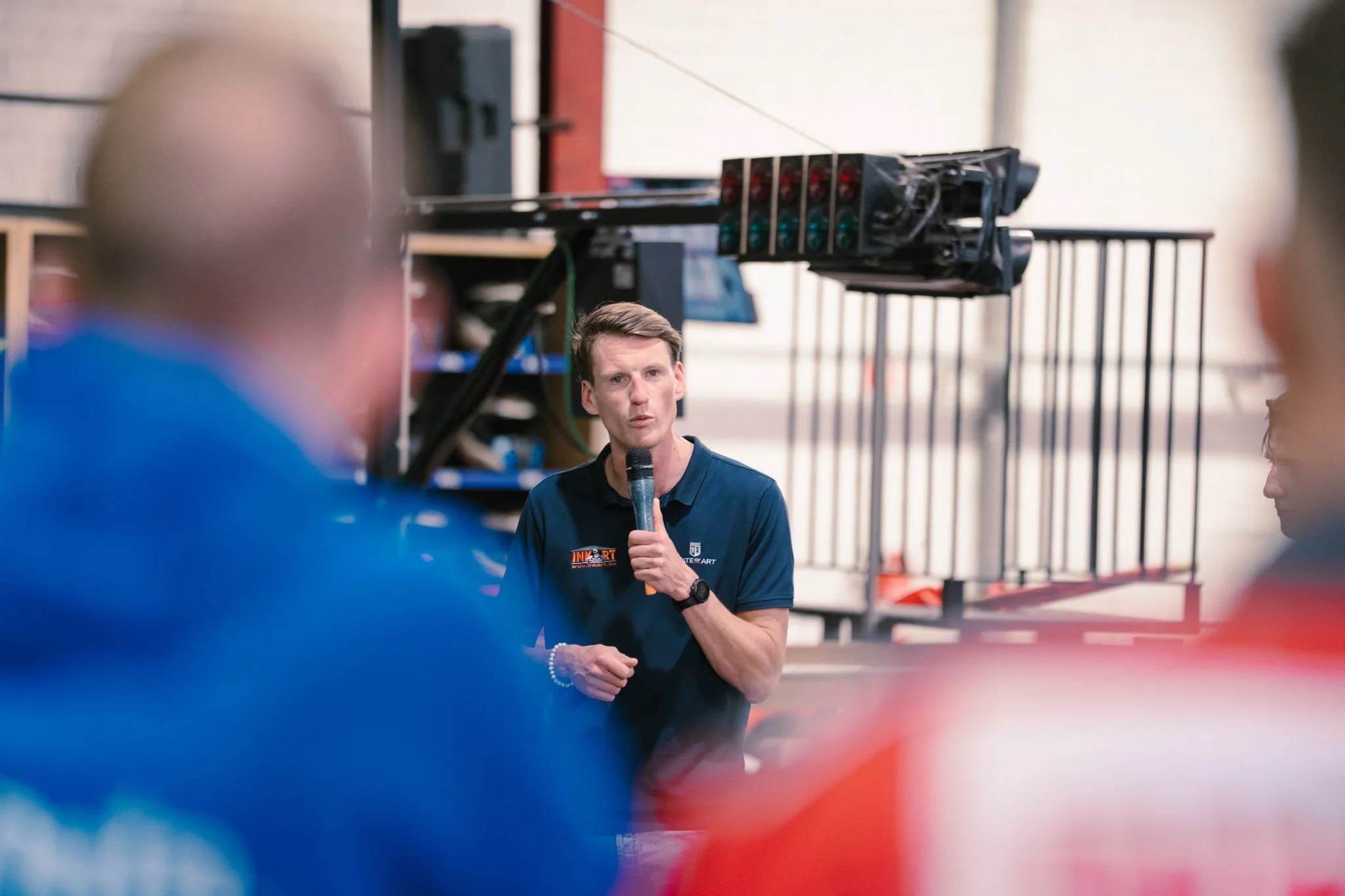 A man holding a microphone speaks in front of a group, with blurred figures in the foreground, in an indoor setting with equipment and a metal railing visible in the background.
