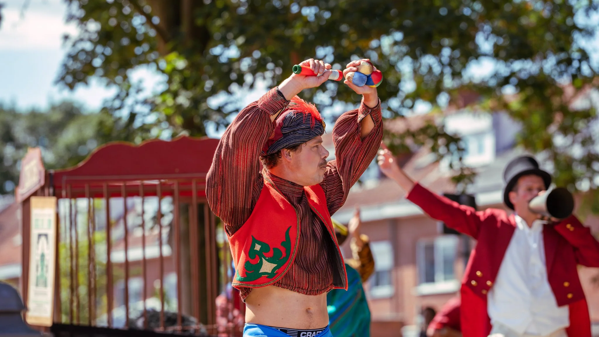 A street performer with red hair and a mustache, wearing a red vest with green designs, is holding a juggling club above his head during a outdoor event. In the background, a man dressed in a red coat and black top hat is playing a horn, and there ar