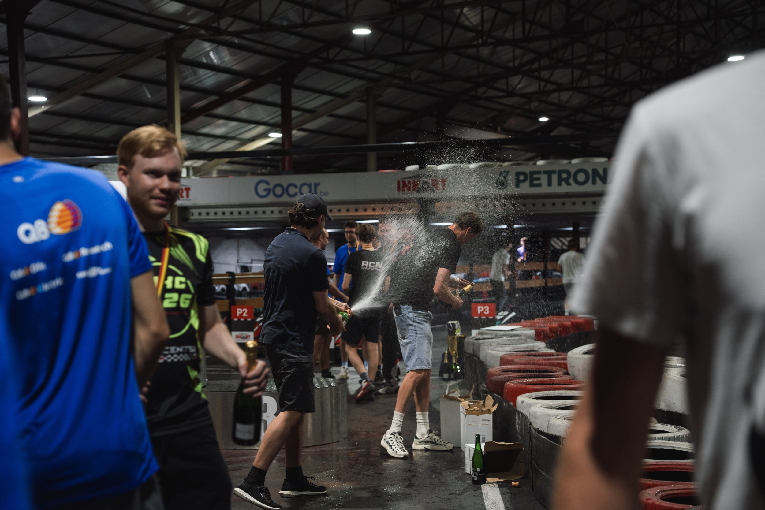 People celebrating with champagne in a garage or pit lane, with a woman spraying champagne and others holding bottles, in a racing environment.