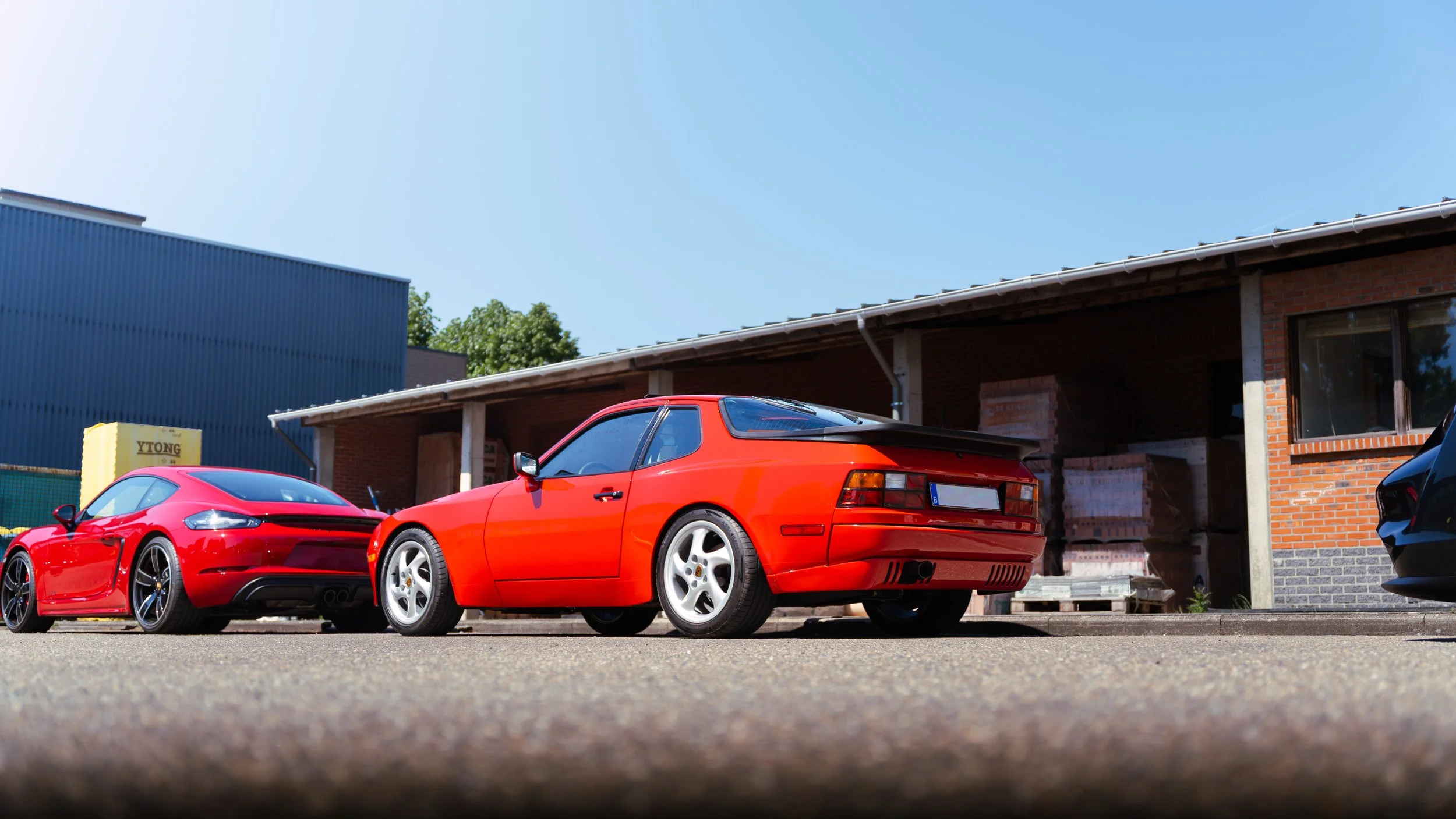 Red sports car and a classic red coupe parked outdoors with a warehouse building in the background.