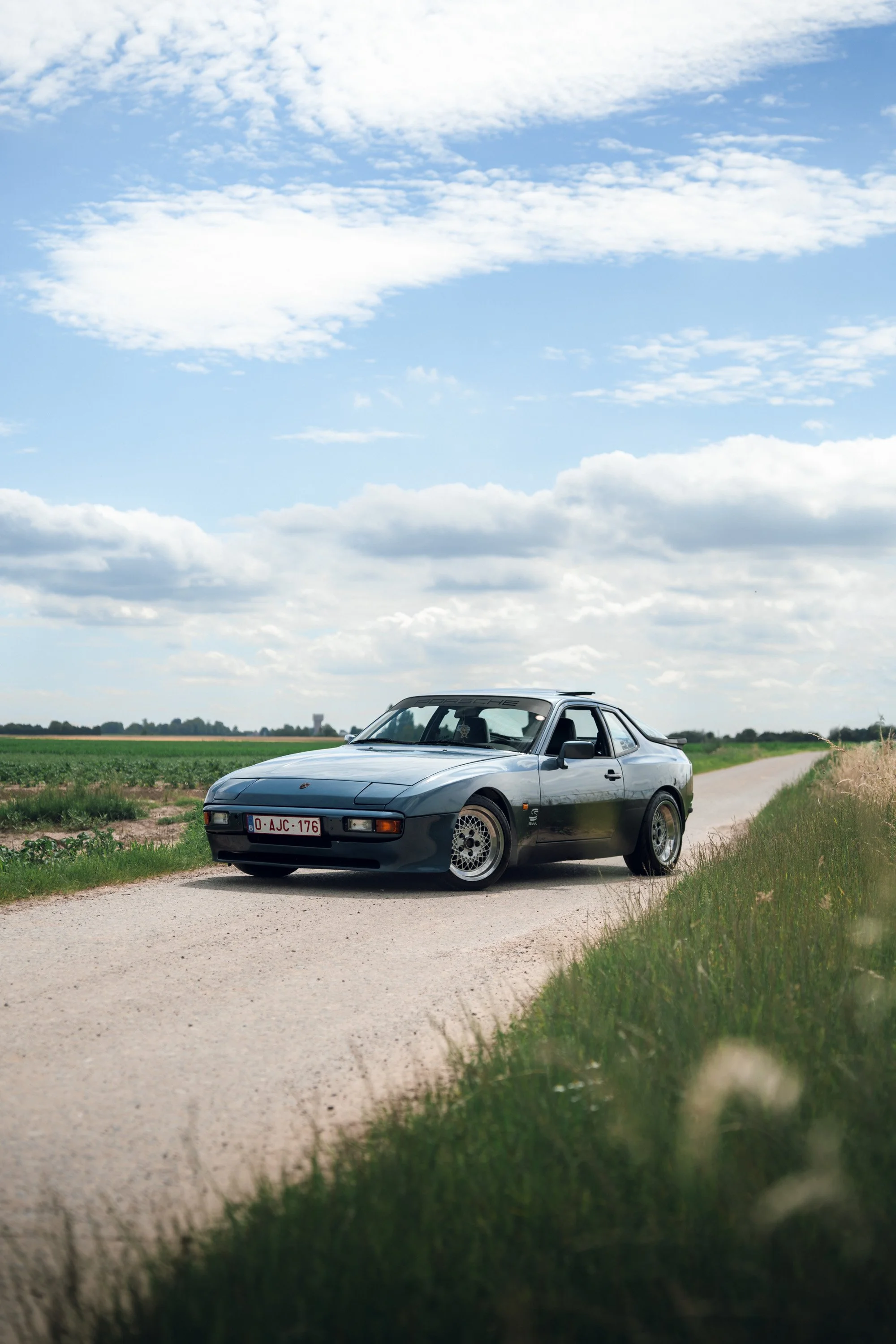 A silver Porsche sports car parked on a dirt road next to a grassy field under a partly cloudy blue sky.