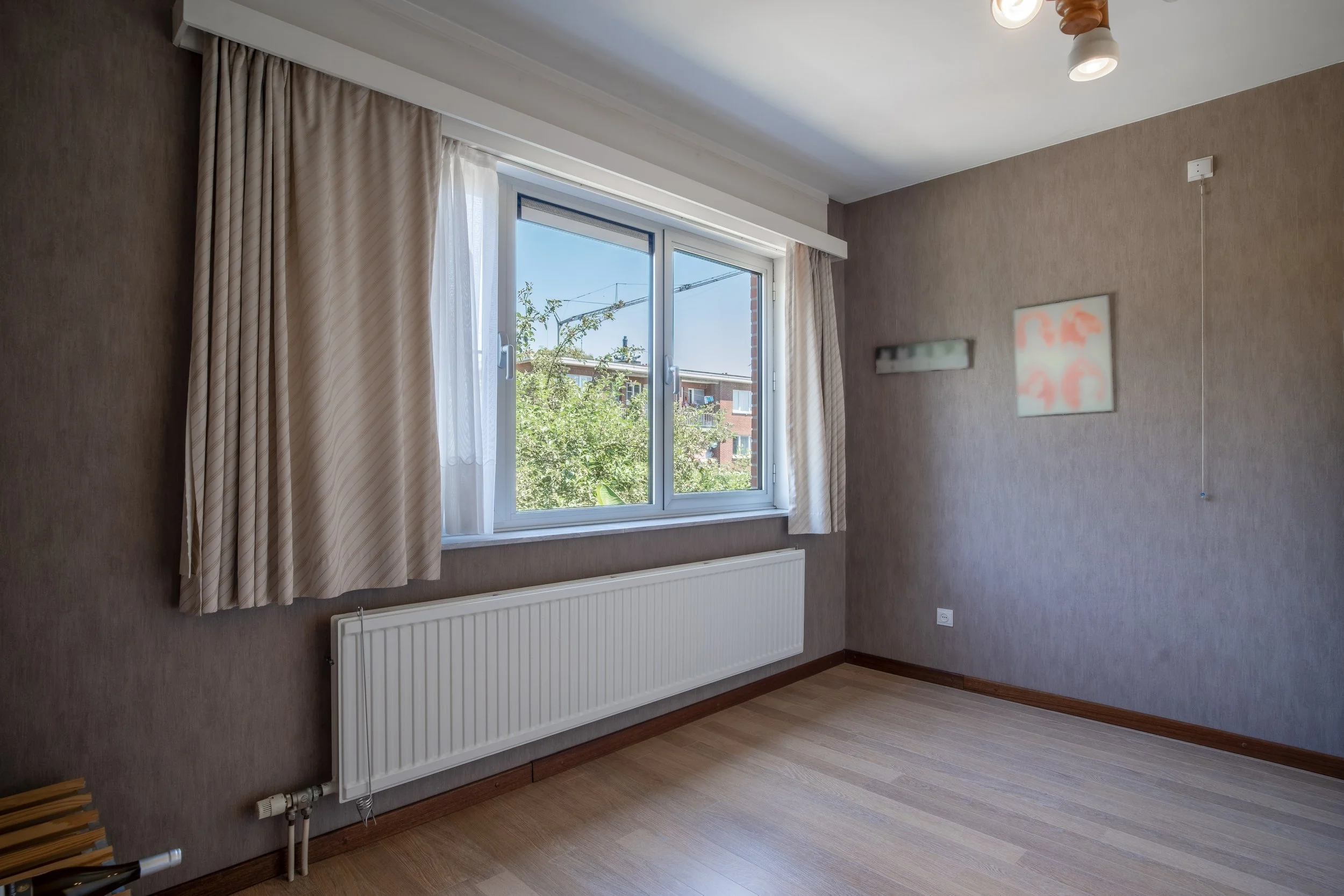 A room with a window, beige curtains, a radiator underneath, and a wall with a piece of blurred artwork, a light fixture, and an electrical socket.