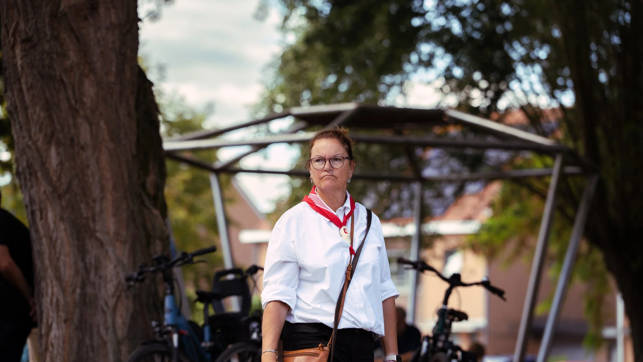 A woman with glasses and a red scarf around her neck, standing outdoors near a tree with a cloudy sky. There are bicycles and a bike rack in the background.