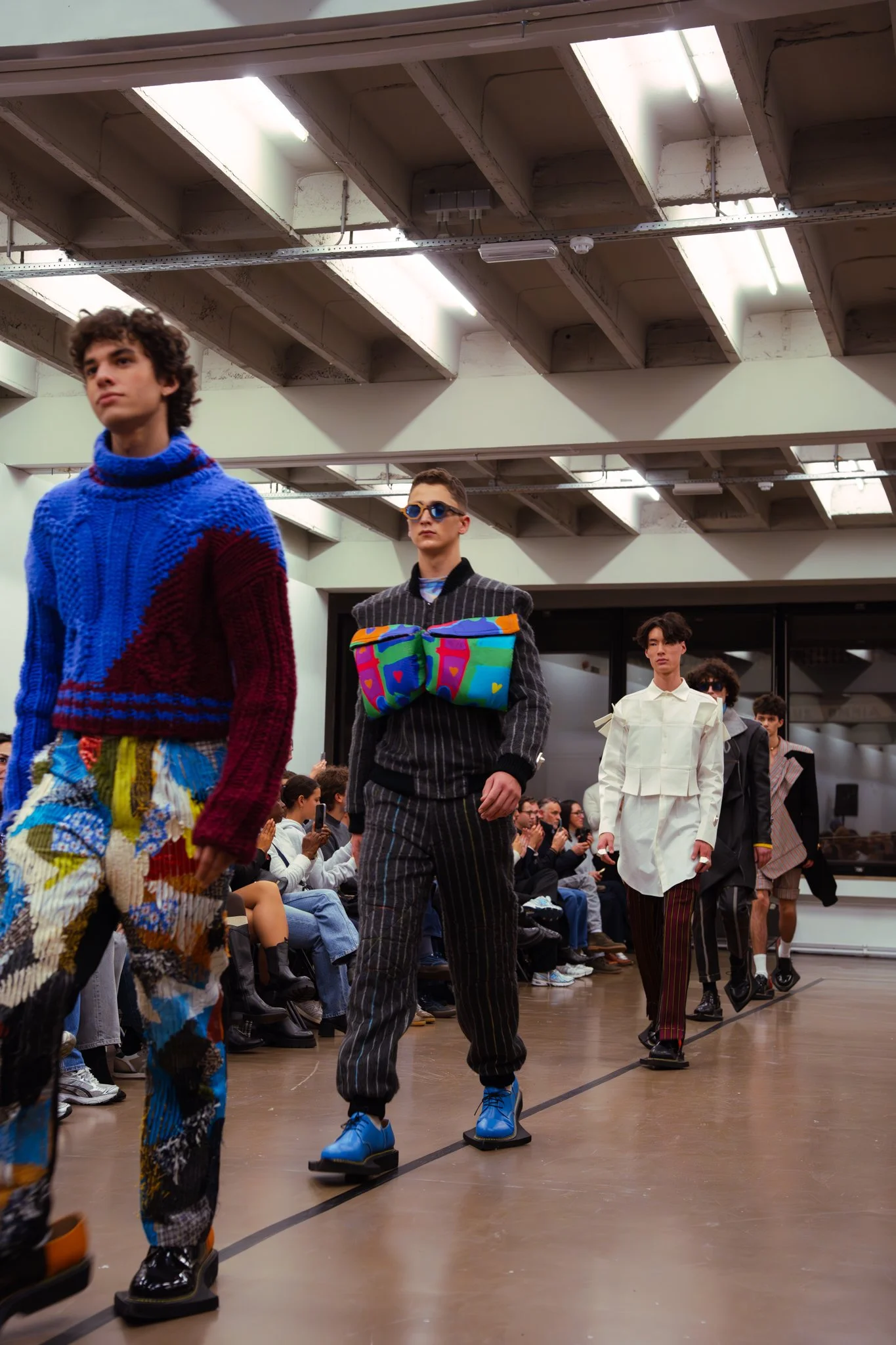 Models walking on a runway during a fashion show, with an audience seated on either side, in an indoor space with skylights overhead.