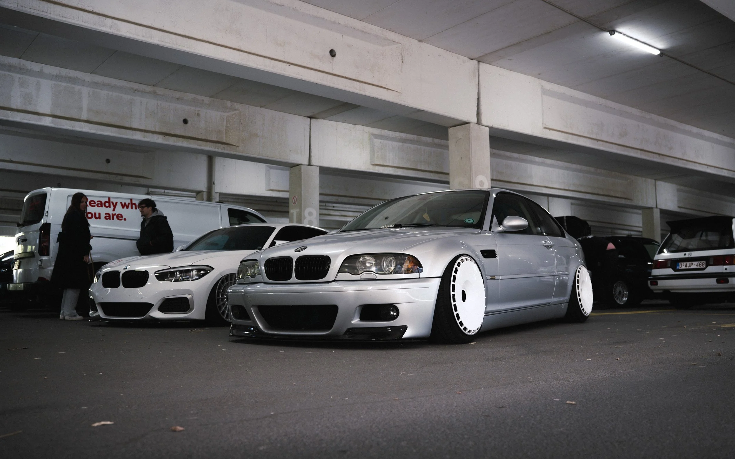 Underground parking garage with several parked cars, including a modified silver BMW Z3 with white wheel covers, and two people standing near a white BMW M4.
