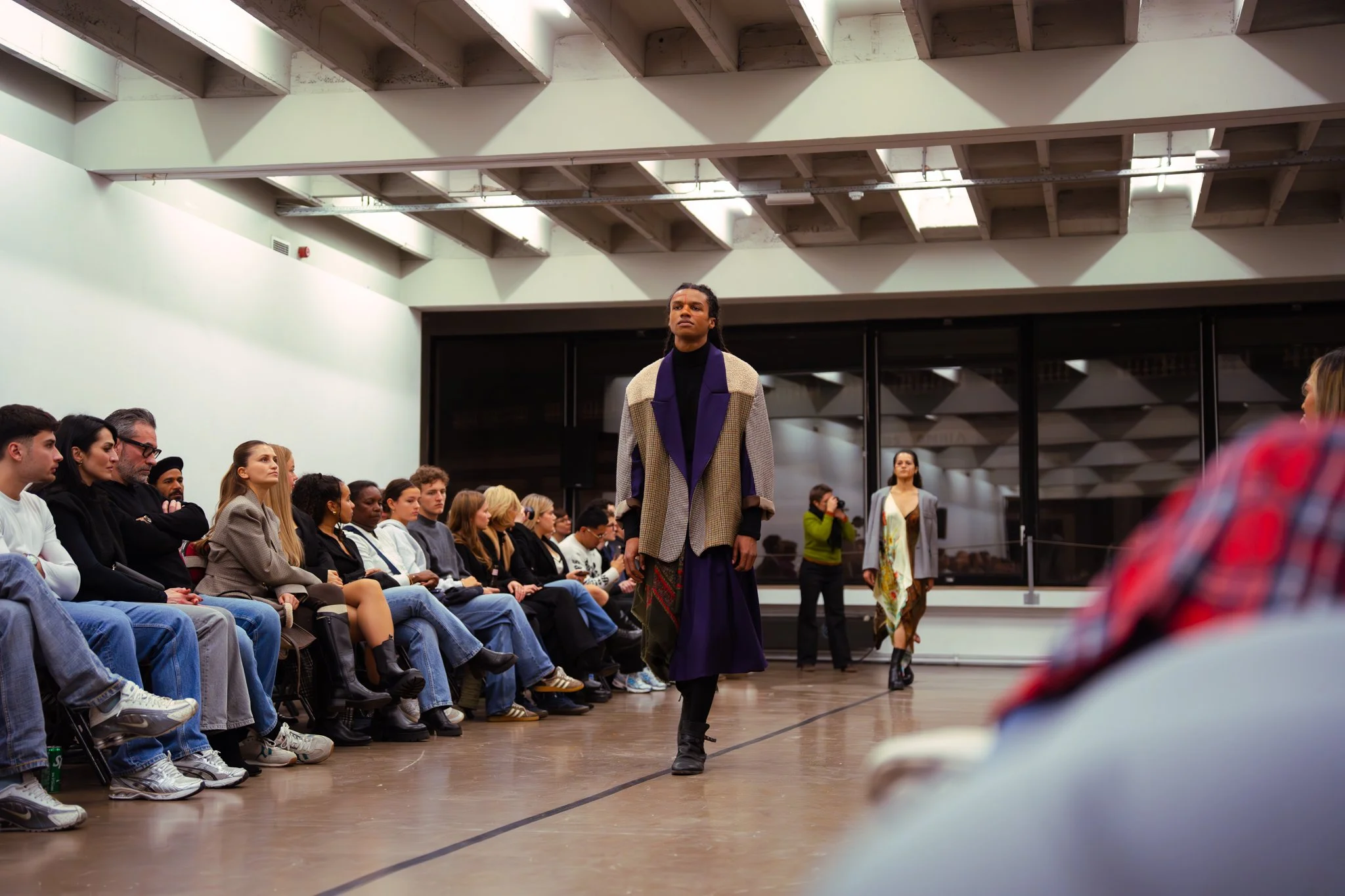 Fashion model walking down runway at a fashion show with audience seated on both sides, and another model in the background.