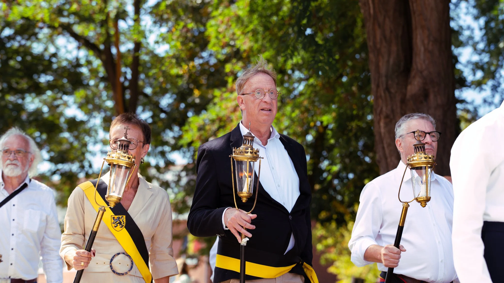 A group of older adults participating in a lantern procession outdoors, holding lanterns during a daytime event with trees in the background.
