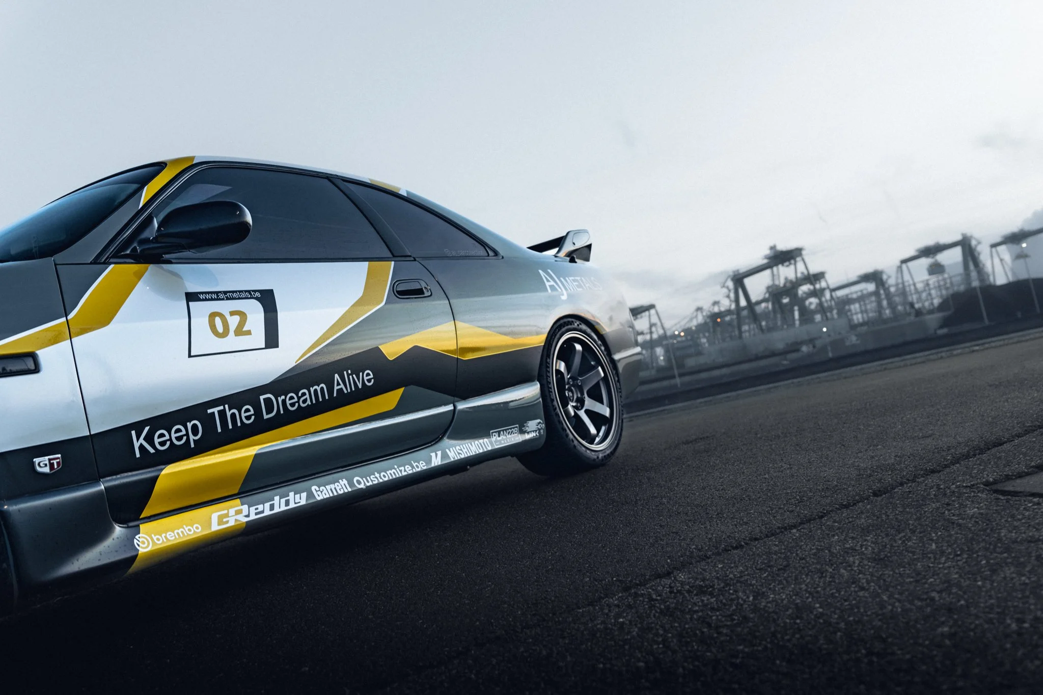 A race car parked on a track with industrial structures in the background. The car has a black, white, and yellow design with various sponsor logos and the phrase 'Keep The Dream Alive' on the side.