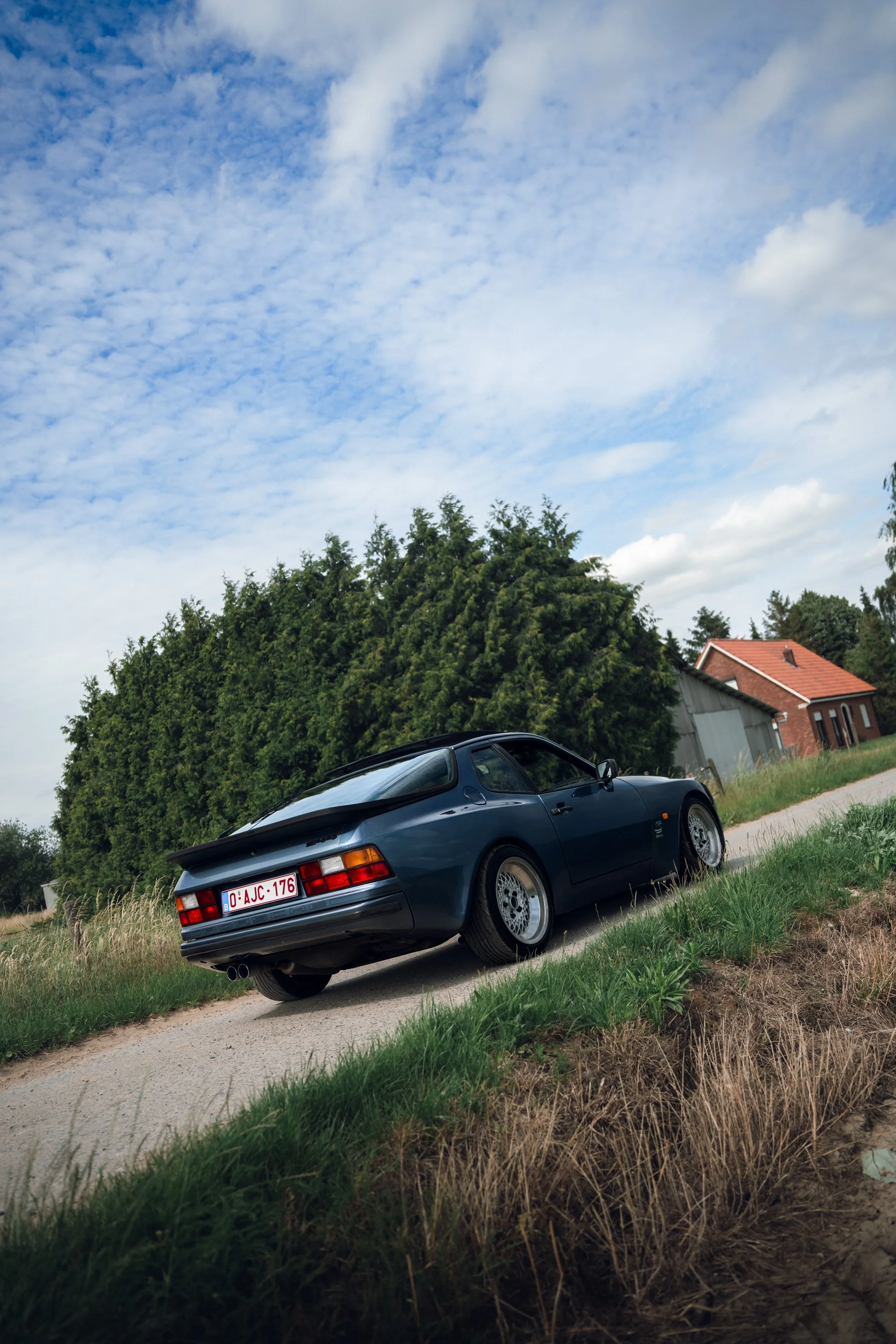 A dark blue vintage sports car parked on a grassy area near trees and houses, with a cloudy sky overhead.