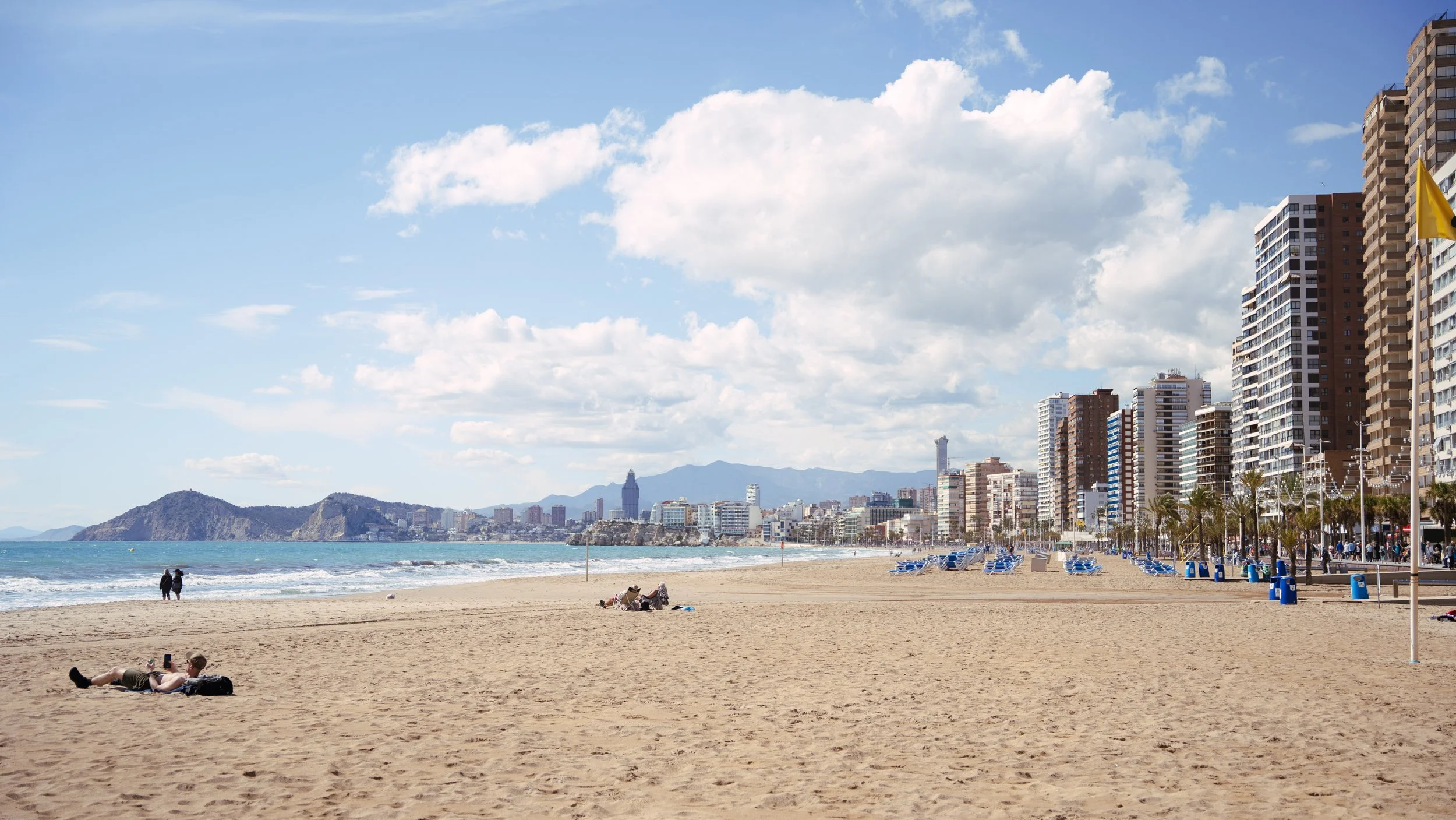 Sunny beach with golden sand, a few people relaxing, and others walking near the shoreline. Tall buildings line the beachfront, with mountains and a city skyline in the background. The sky is mostly clear with some clouds.