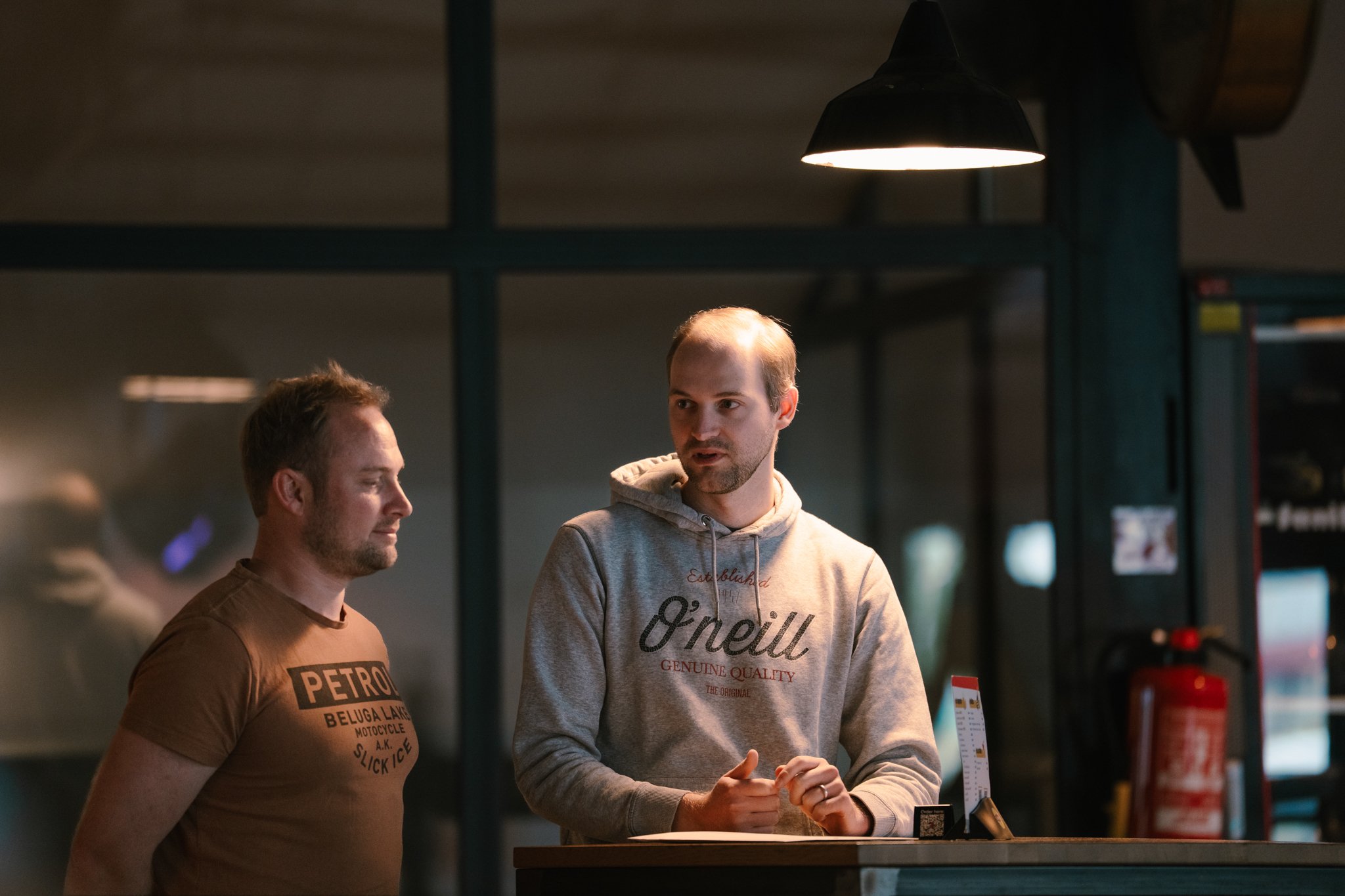 Two men talking in an indoor setting, one wearing a brown T-shirt and the other a gray O'Neill hoodie, illuminated by overhead lighting.