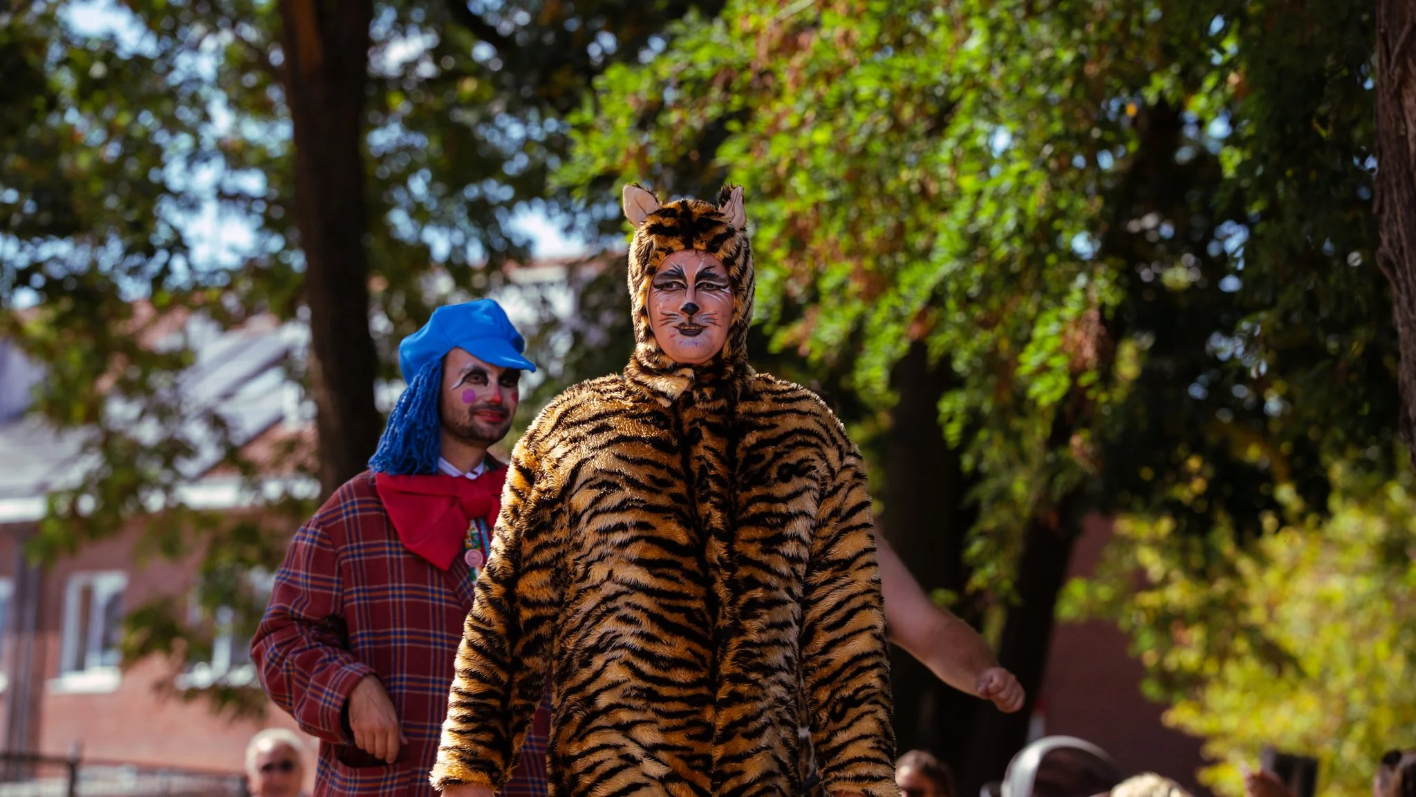 People dressed as clowns and animals participating in a parade or festival outdoors during the day, surrounded by trees and buildings.