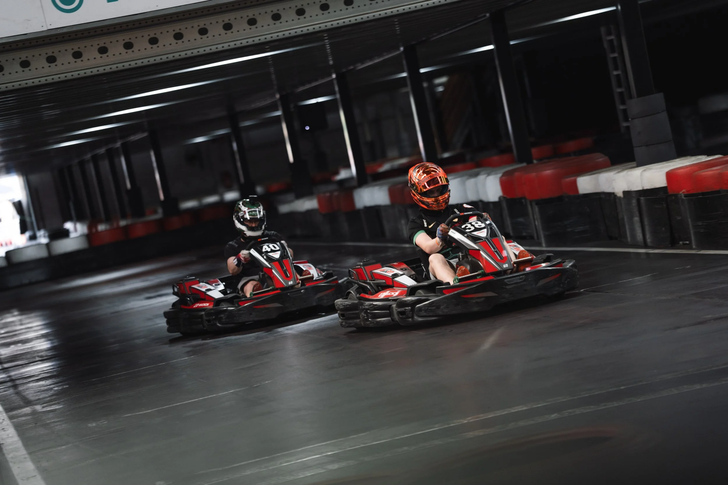 Two go-karts with riders racing on an indoor track under a metal ceiling.