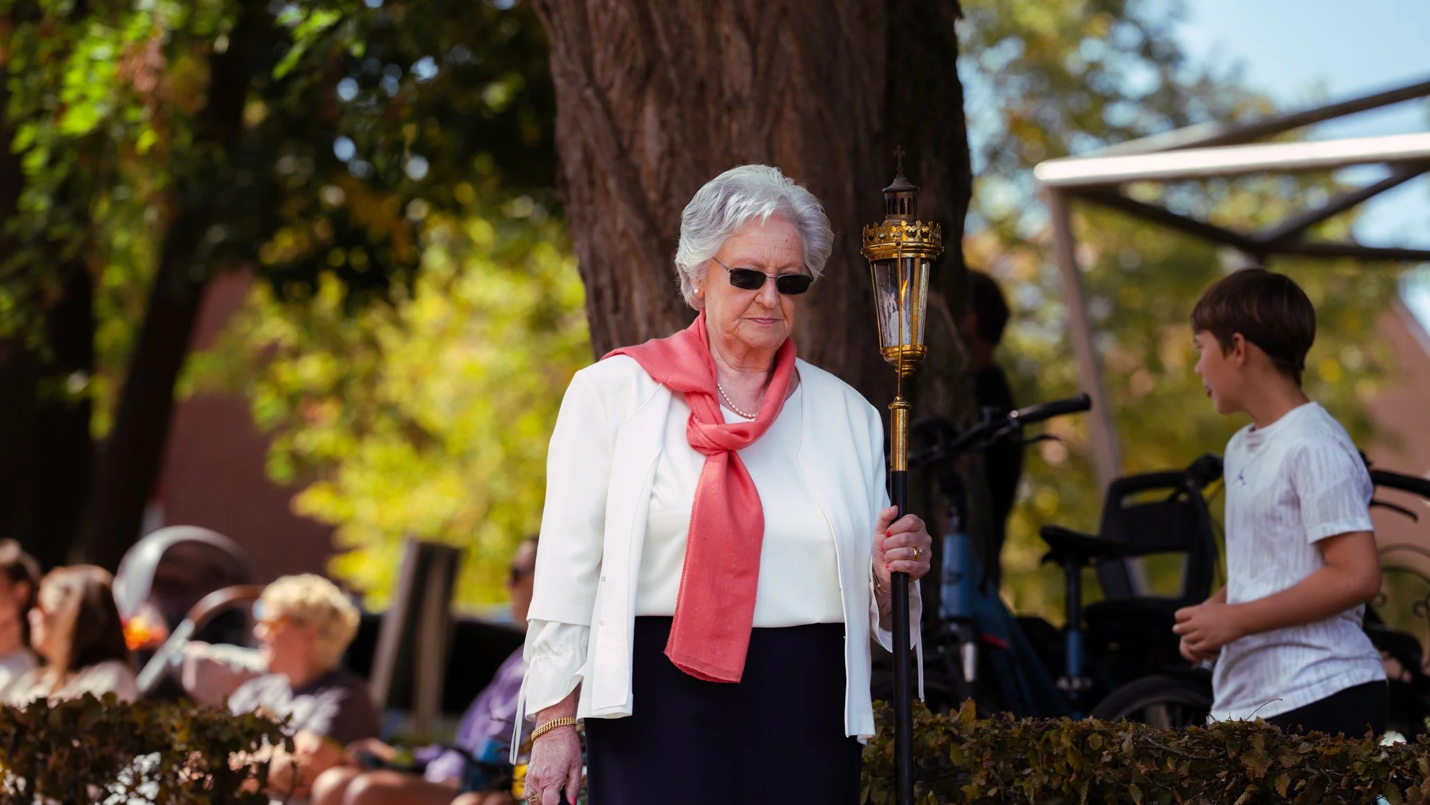 An elderly woman with white hair, wearing sunglasses, a white jacket, and a peach-colored scarf, holding a liturgical candle stand, standing outdoors in a park with trees and other people seated in the background.
