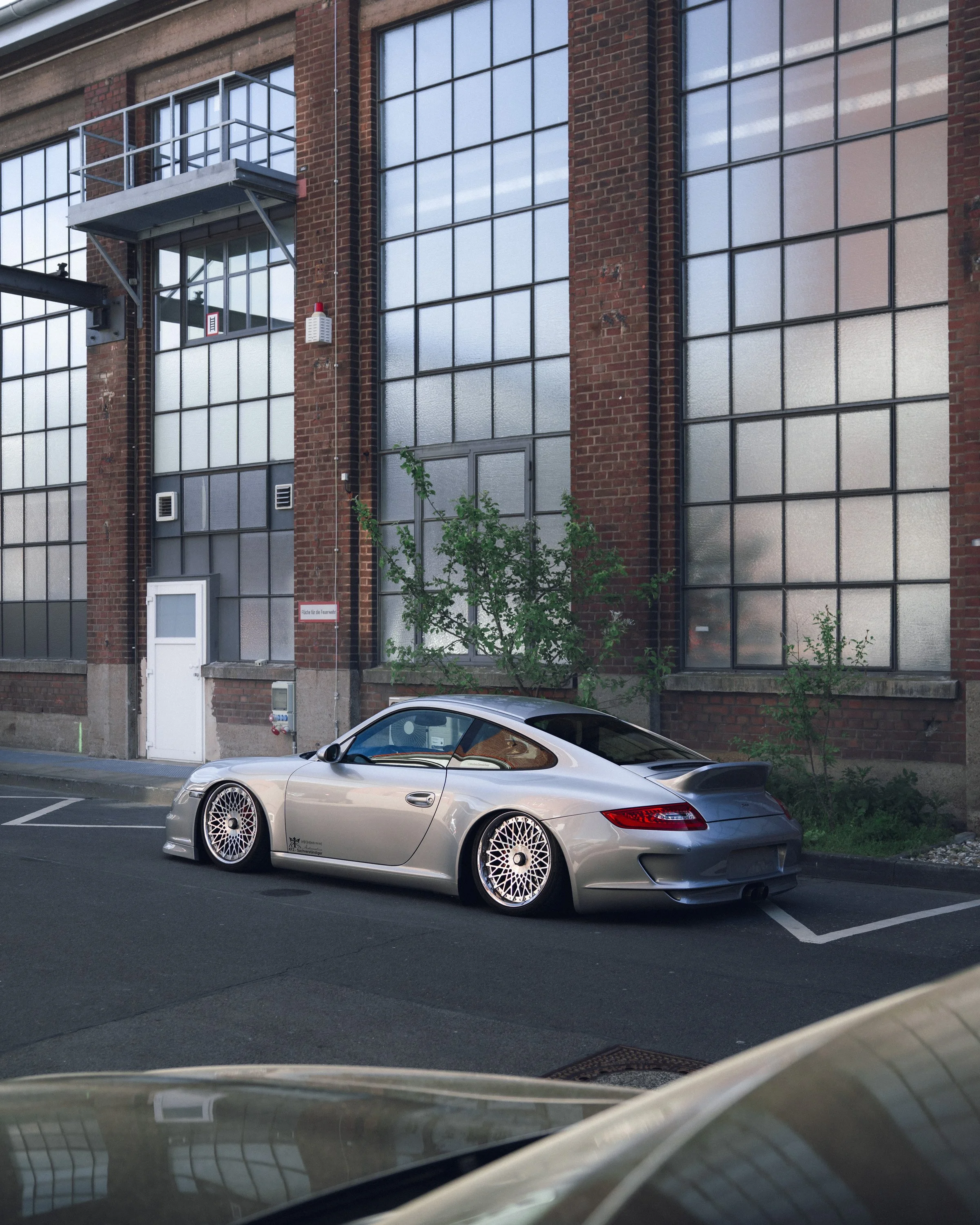 A silver sports car with customized white wire wheels parked in a parking lot in front of a red brick industrial building with large glass windows.