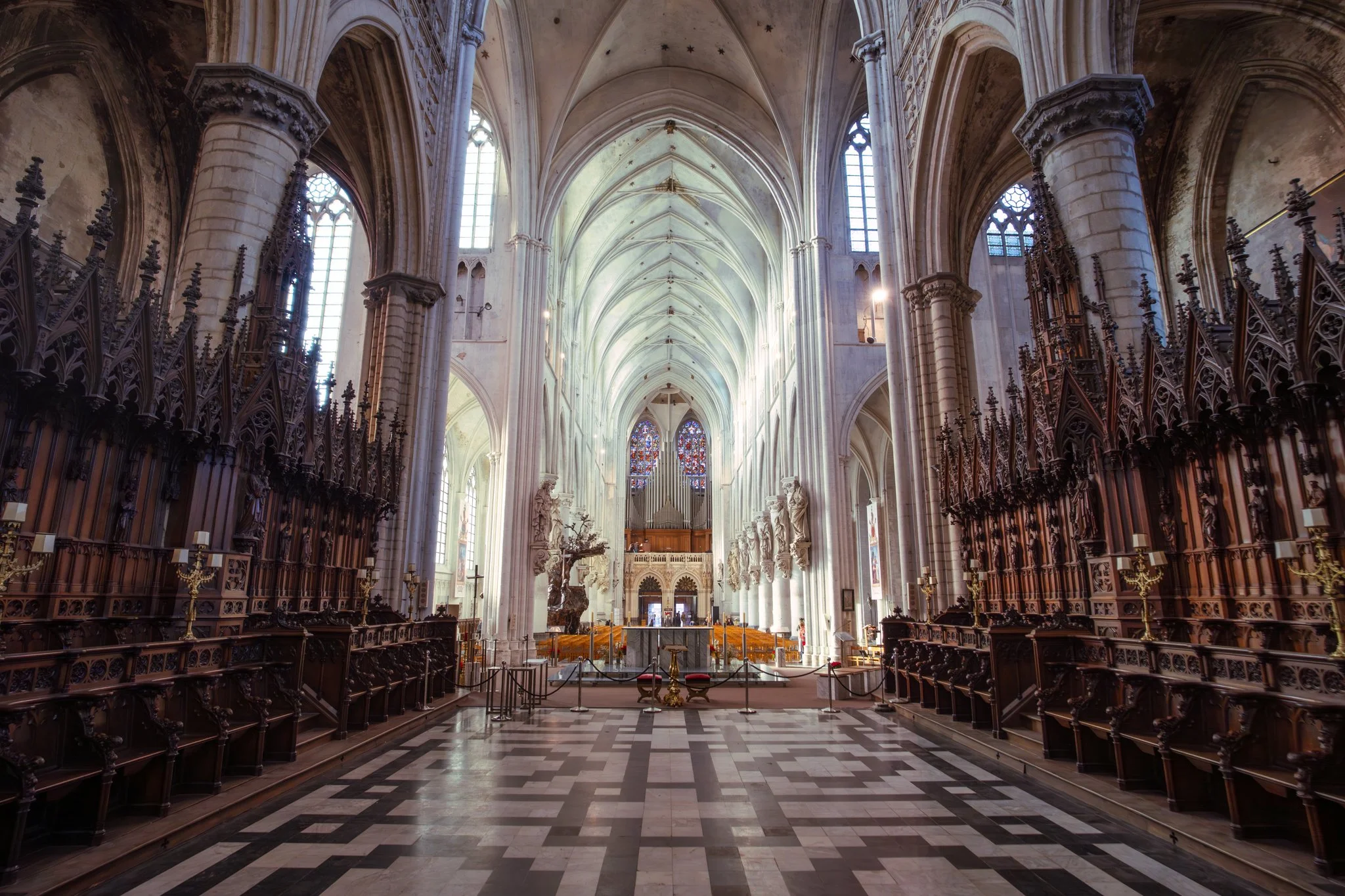 Interior view of a Gothic cathedral with high vaulted ceilings, stained glass windows, and wooden pews on both sides of the aisle.
