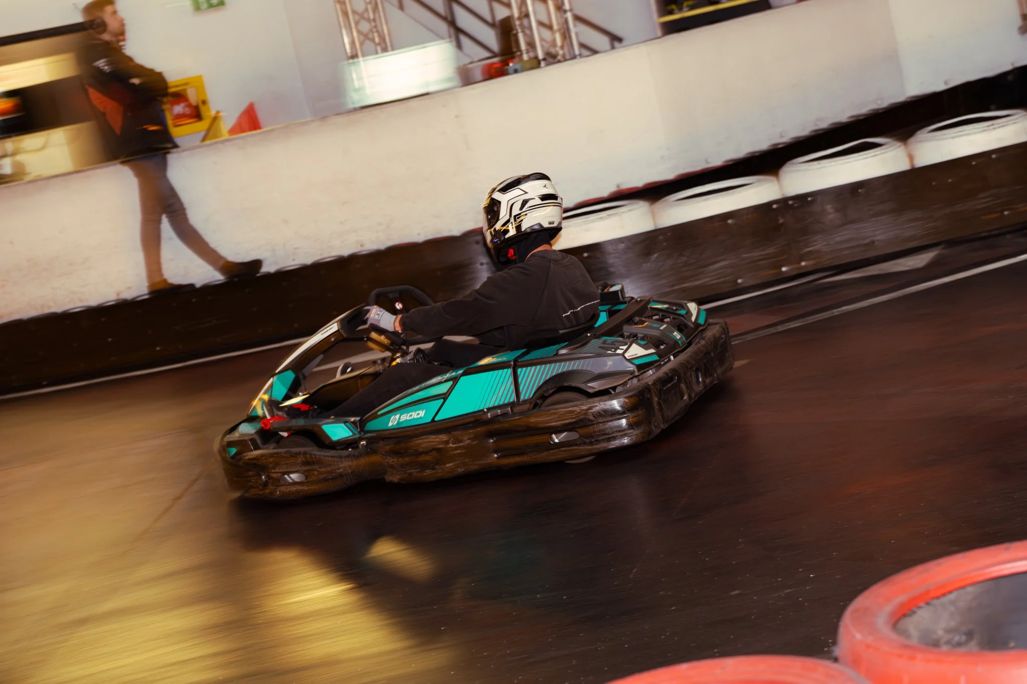 A person wearing a helmet riding a black and turquoise electric go-kart on an indoor track with a shiny, dark floor. A reflective wall shows a blurred figure walking by.