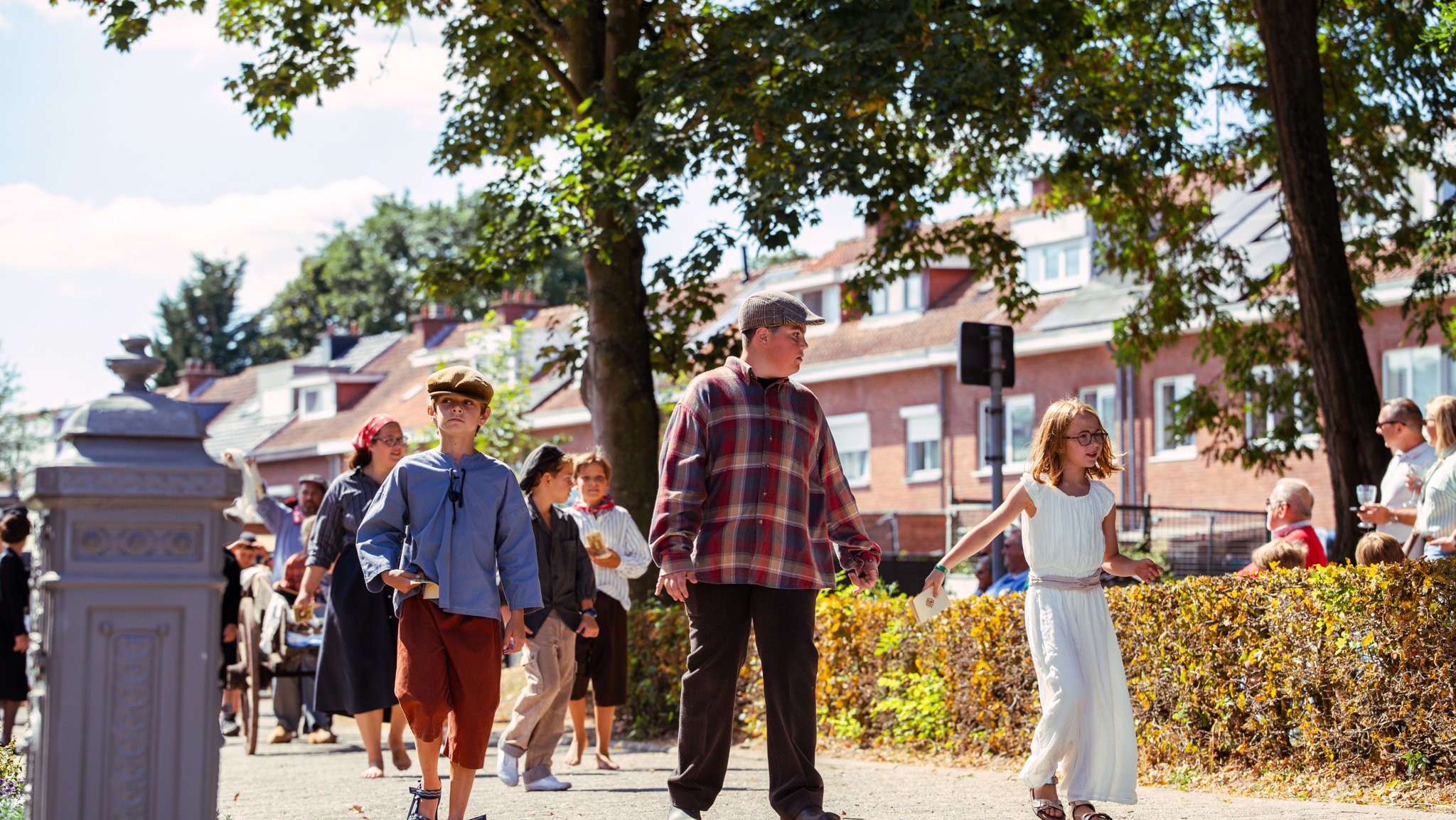 People dressed in vintage clothing walking outdoors on a sunny day, with trees and buildings in the background.