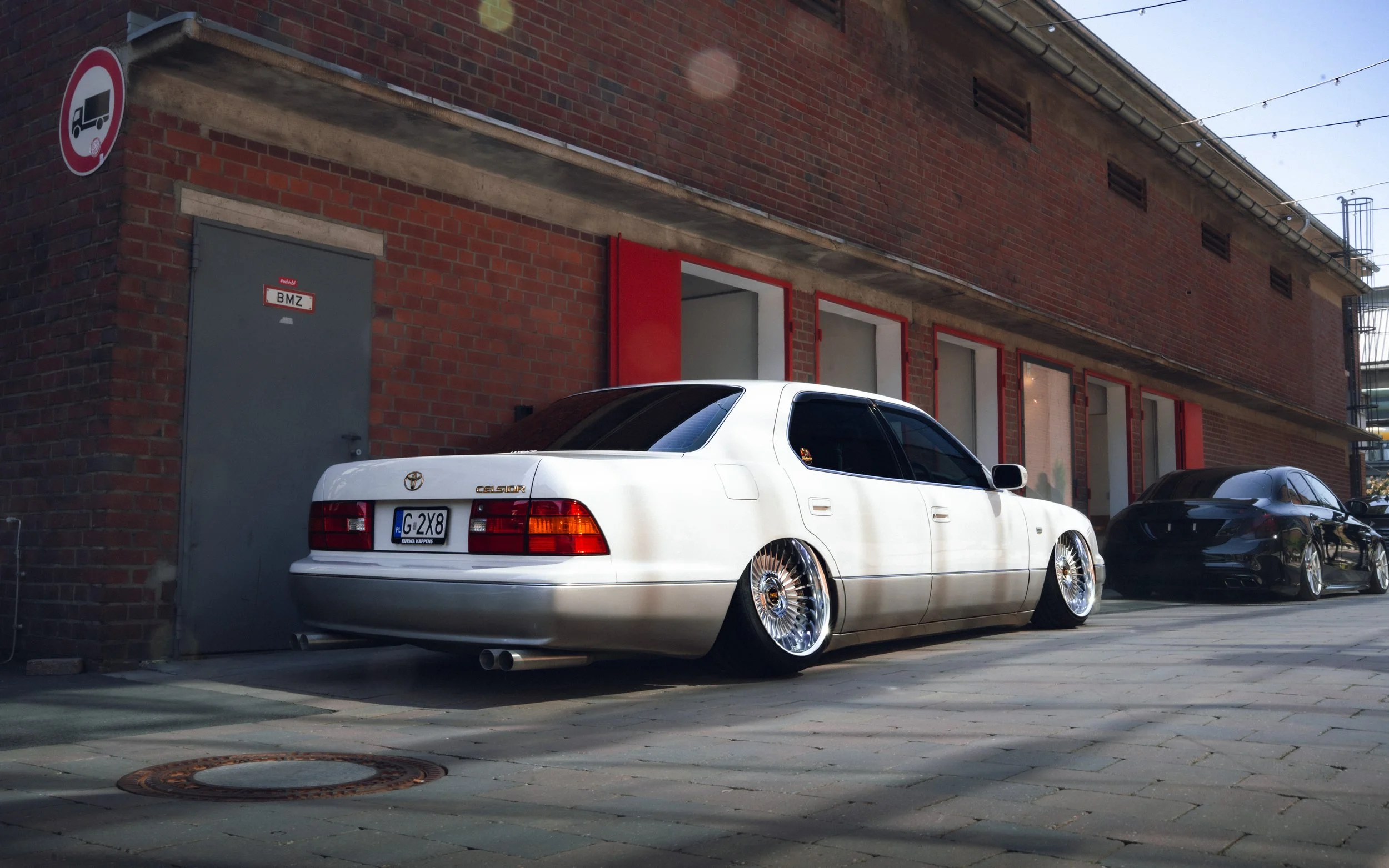 A white Toyota Celsior car with custom rims parked beside a brick building with large windows and red framing, under a clear sky.