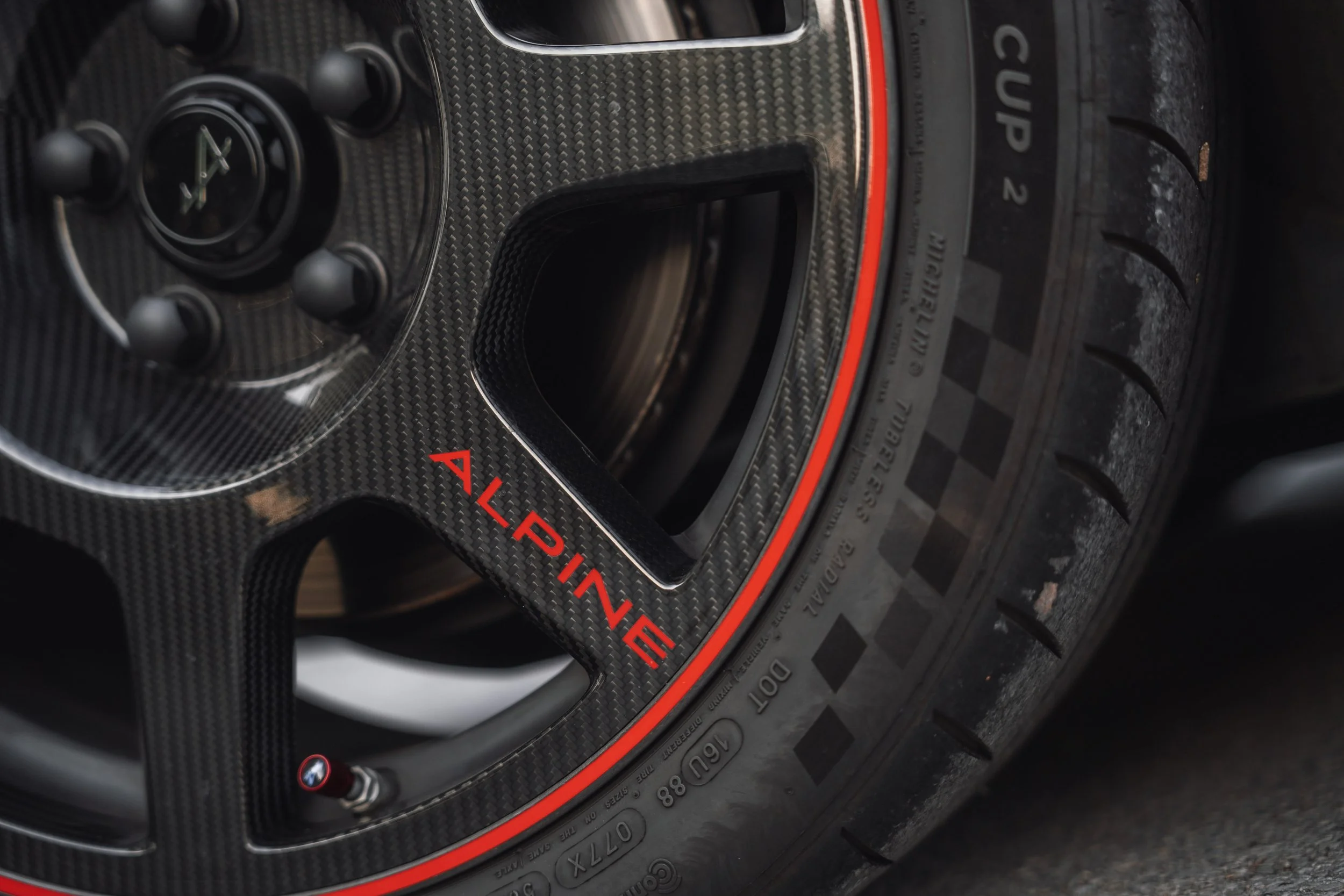 Close-up of a racing car tire with a carbon fiber rear wing and a tire pressure warning label. The focus is on the intricate details of the carbon fiber, the tire tread, and a warning message that reads 'CHECK PRESSURE' in red letters.
