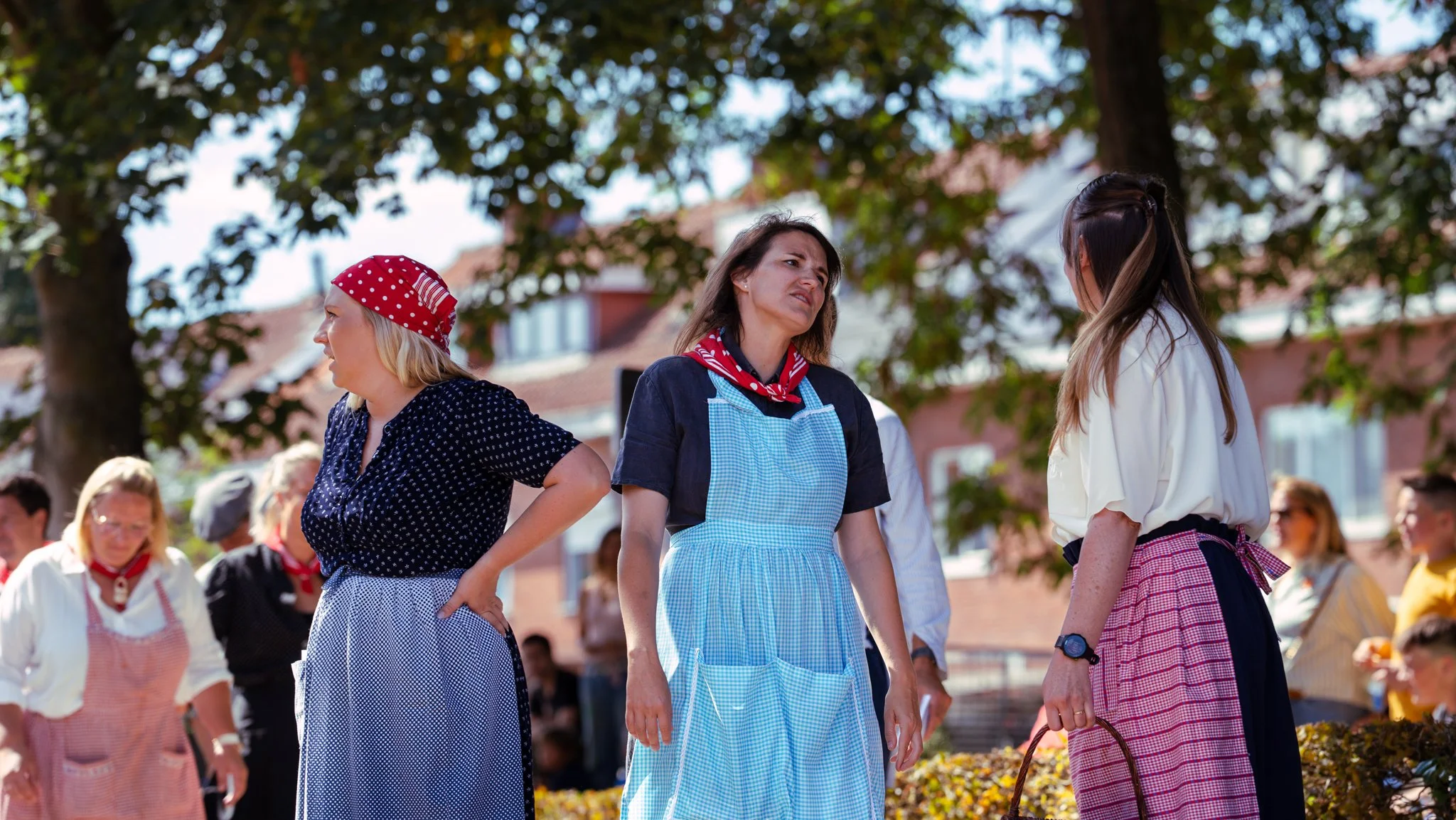 Three women dressed in traditional or work attire engaged in a conversation outdoors, with other people and trees in the background.
