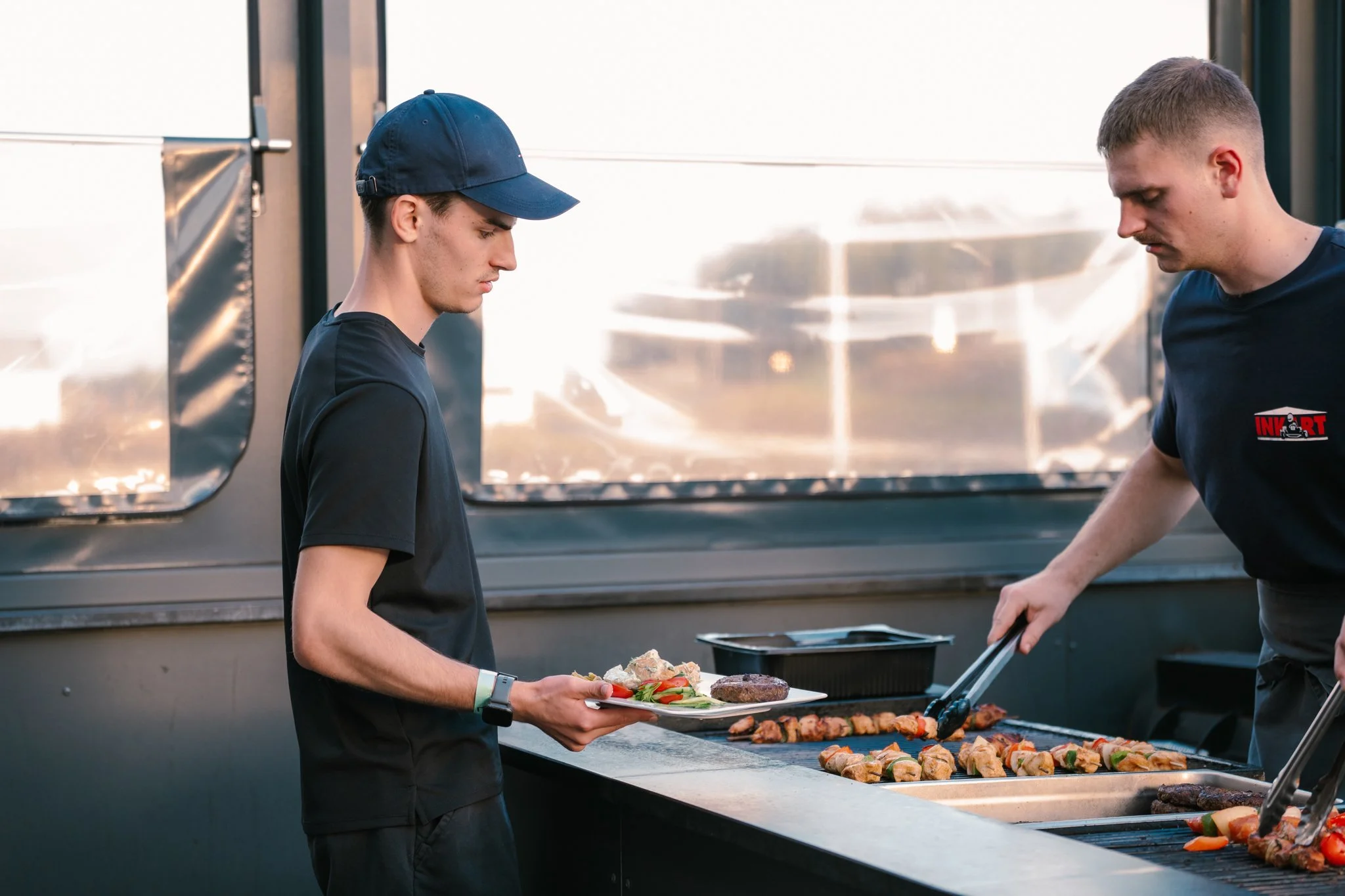 Two young men are grilling food outdoors, one is serving a plate of food while the other is tending to the grill with tongs. The grill has skewers of meat and vegetables.