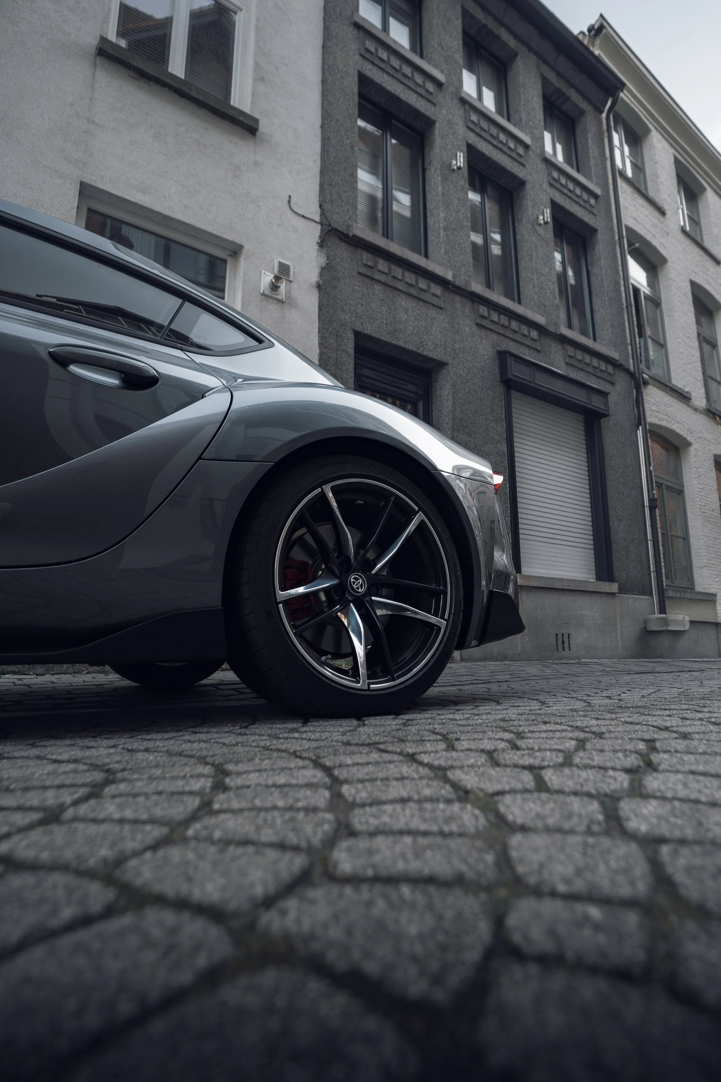 A vintage gray sports car parked on a cobblestone street in front of a multi-story residential building with dark and white facades and large windows.