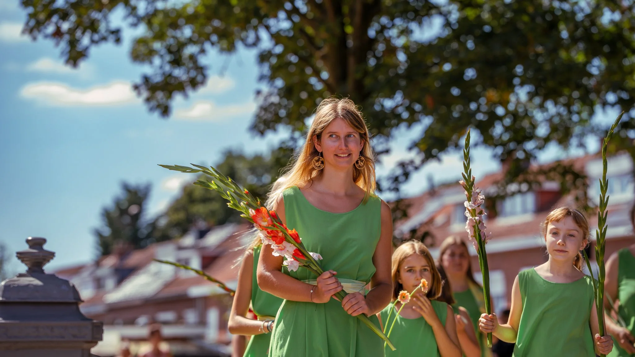 A group of young women and girls in green dresses holding flowers during an outdoor event on a sunny day, with trees and houses in the background.