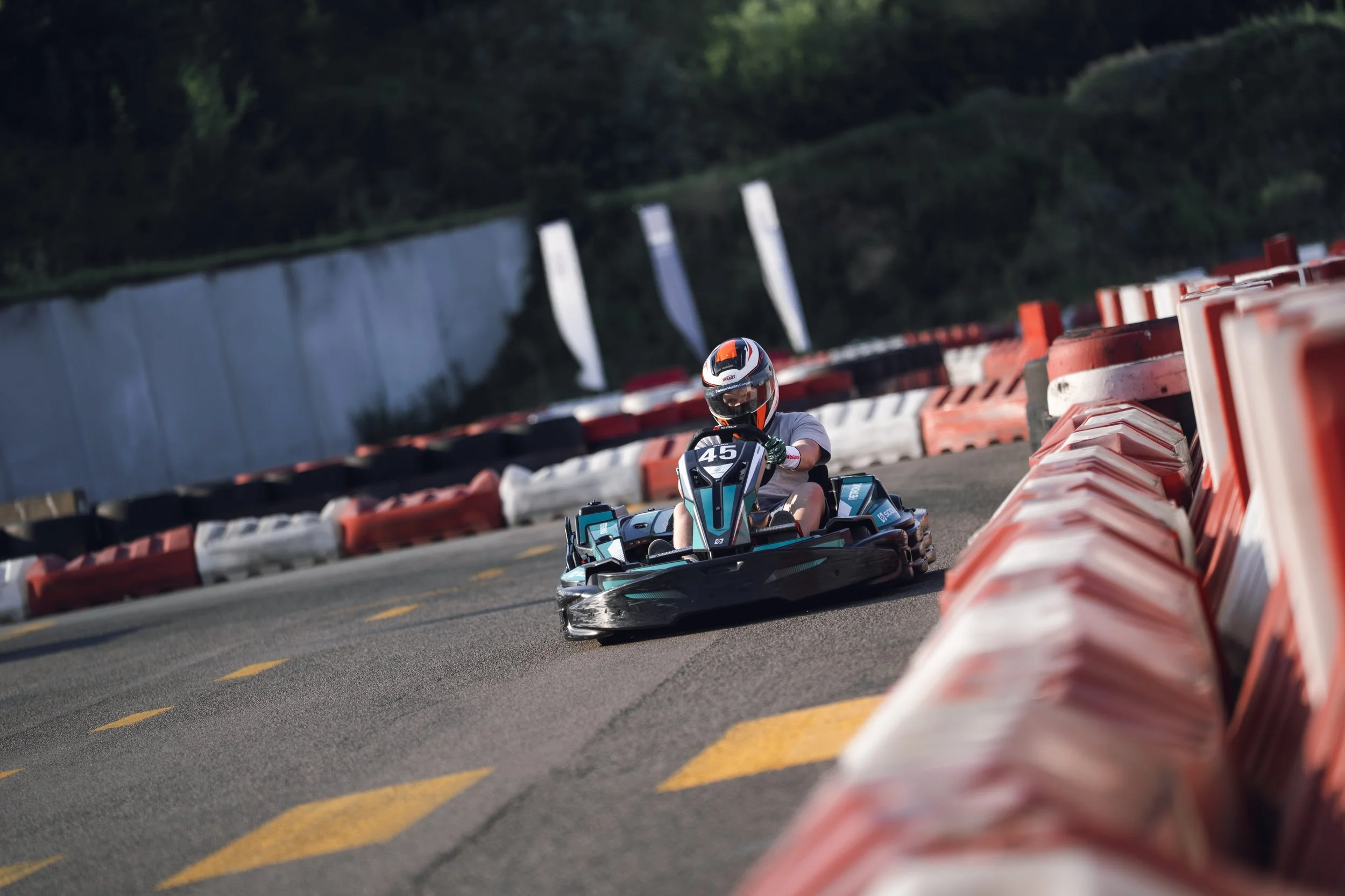 A person riding a go-kart with the number 45, wearing a helmet, on an outdoor race track lined with orange and white barriers, surrounded by green trees.