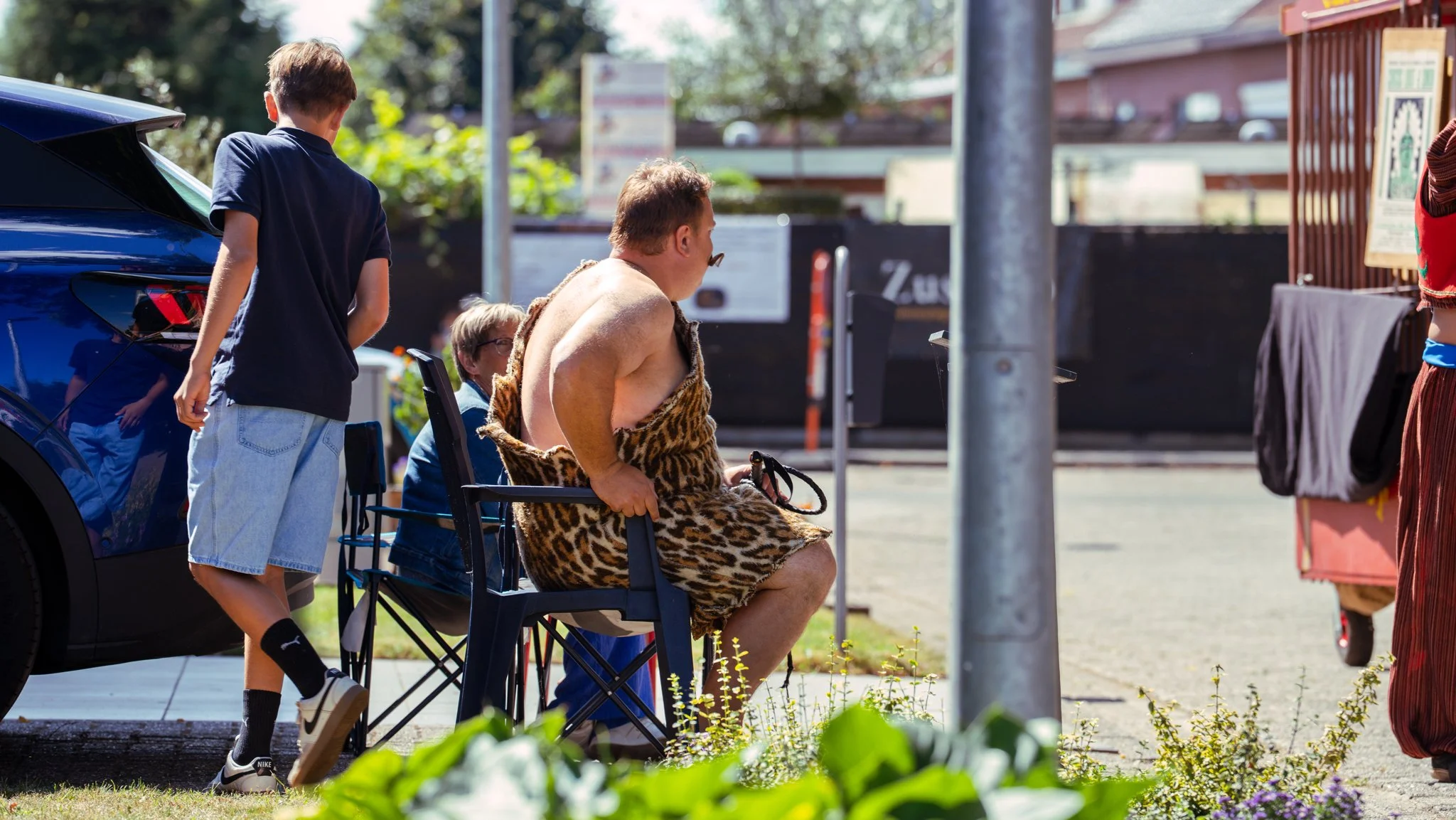 A man wearing a leopard print cloth draped over his lap, sitting on a black chair at an outdoor setting. He appears to be in a humorous situation, with his shirt pulled down, exposing his shoulder and upper body, while he holds a small object in his 