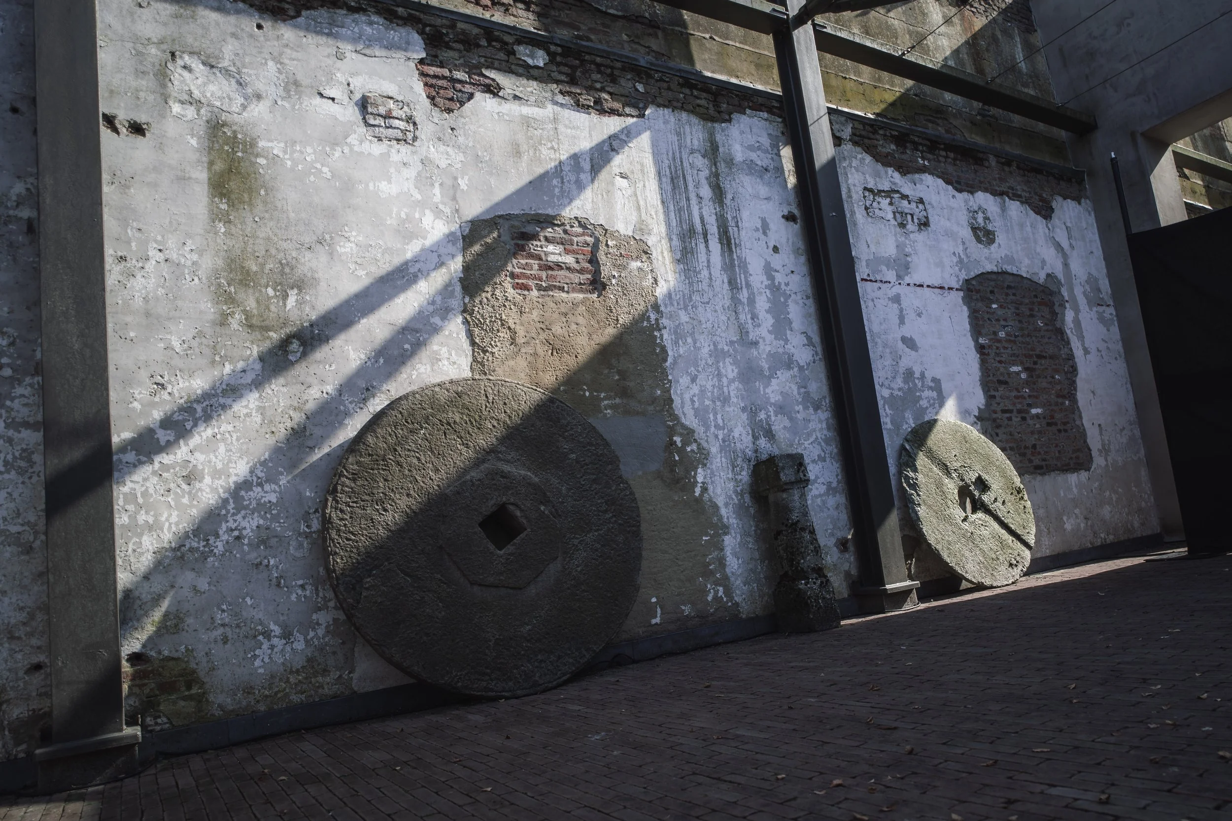 A brick wall with peeling white paint and various circular stone millstones leaning against it. Steel beams frame the scene and shadows fall across the wall and ground, which is paved with bricks.