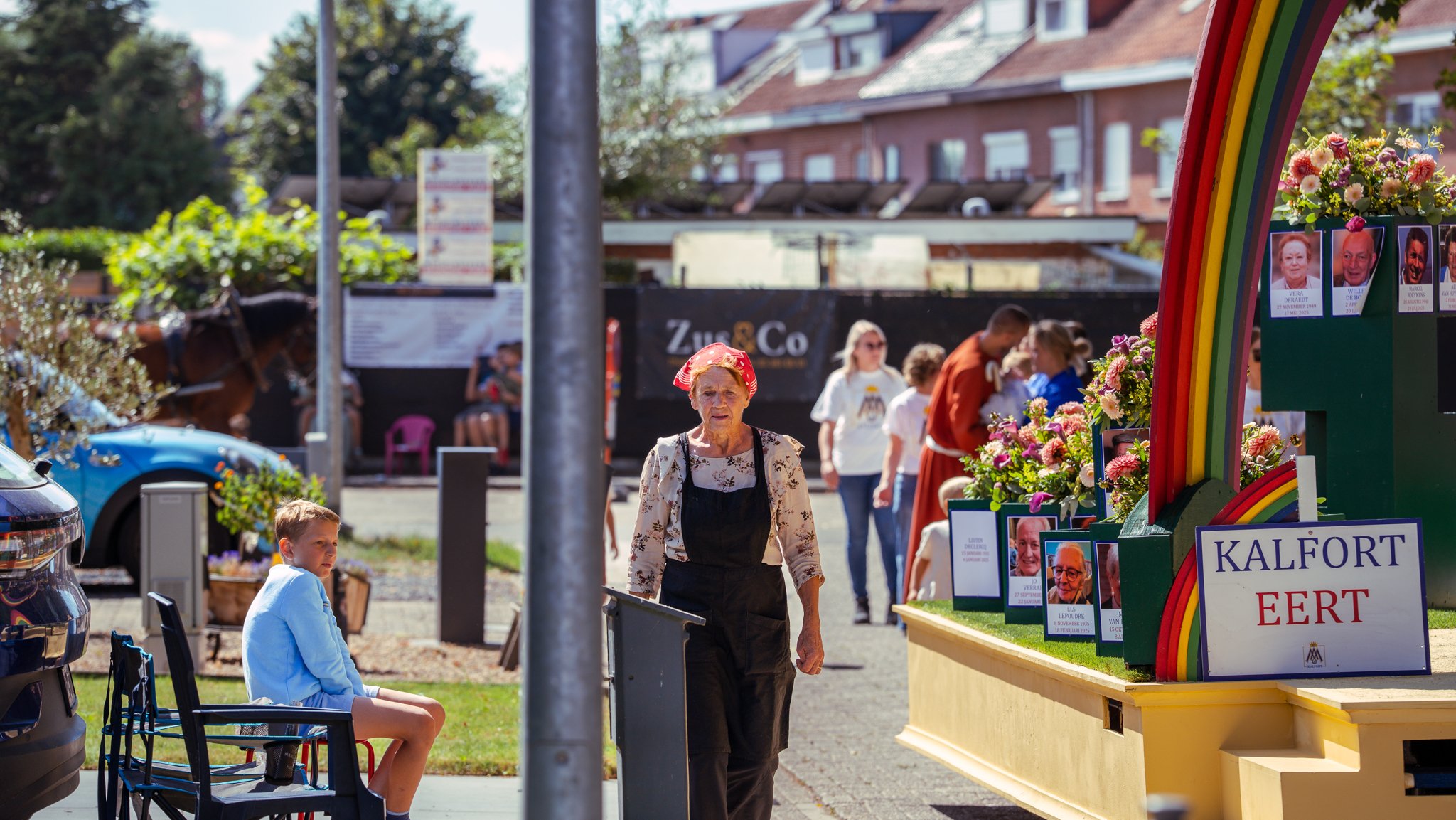 People attending an outdoor event with a colorful display featuring photographs and flowers, including an elderly woman in apron and headscarf, and a young boy seated nearby.