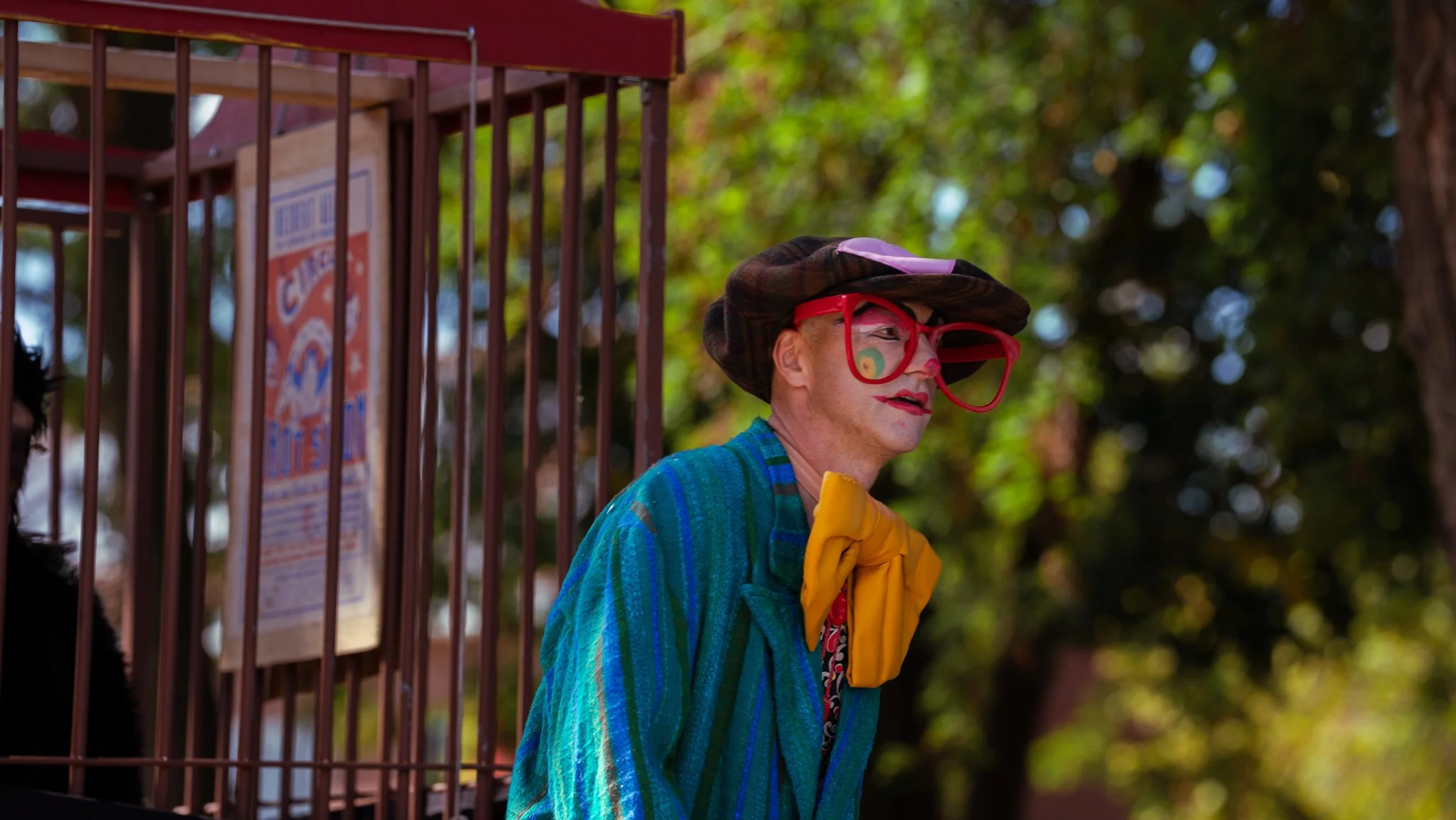 Person dressed as a clown, wearing large red glasses, a wide-brimmed hat, a colorful striped blue jacket, and a yellow bow tie, standing outdoors near a metal cage with trees in the background.