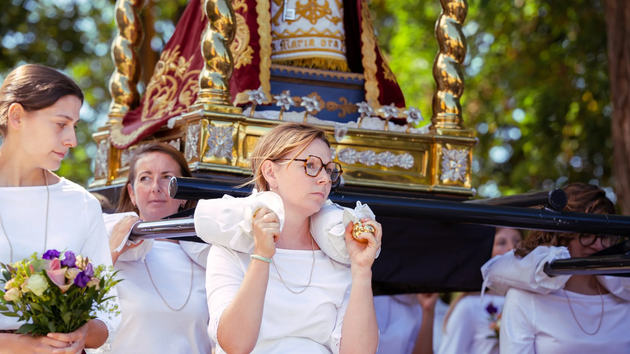 Women participating in a religious procession, carrying a float with a religious figure adorned with gold and red decorations, surrounded by greenery.