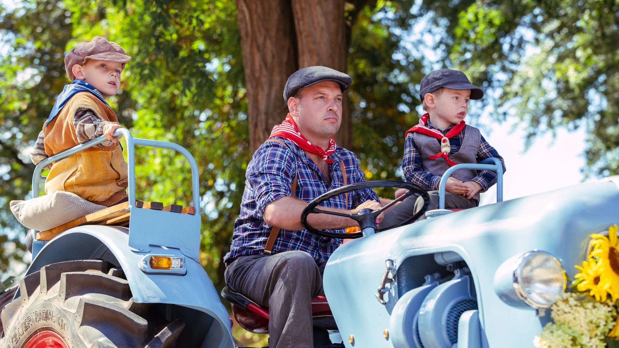 A man with two young boys riding on a vintage blue tractor outdoors, surrounded by trees and sunlight, dressed in old-fashioned clothing and hats.