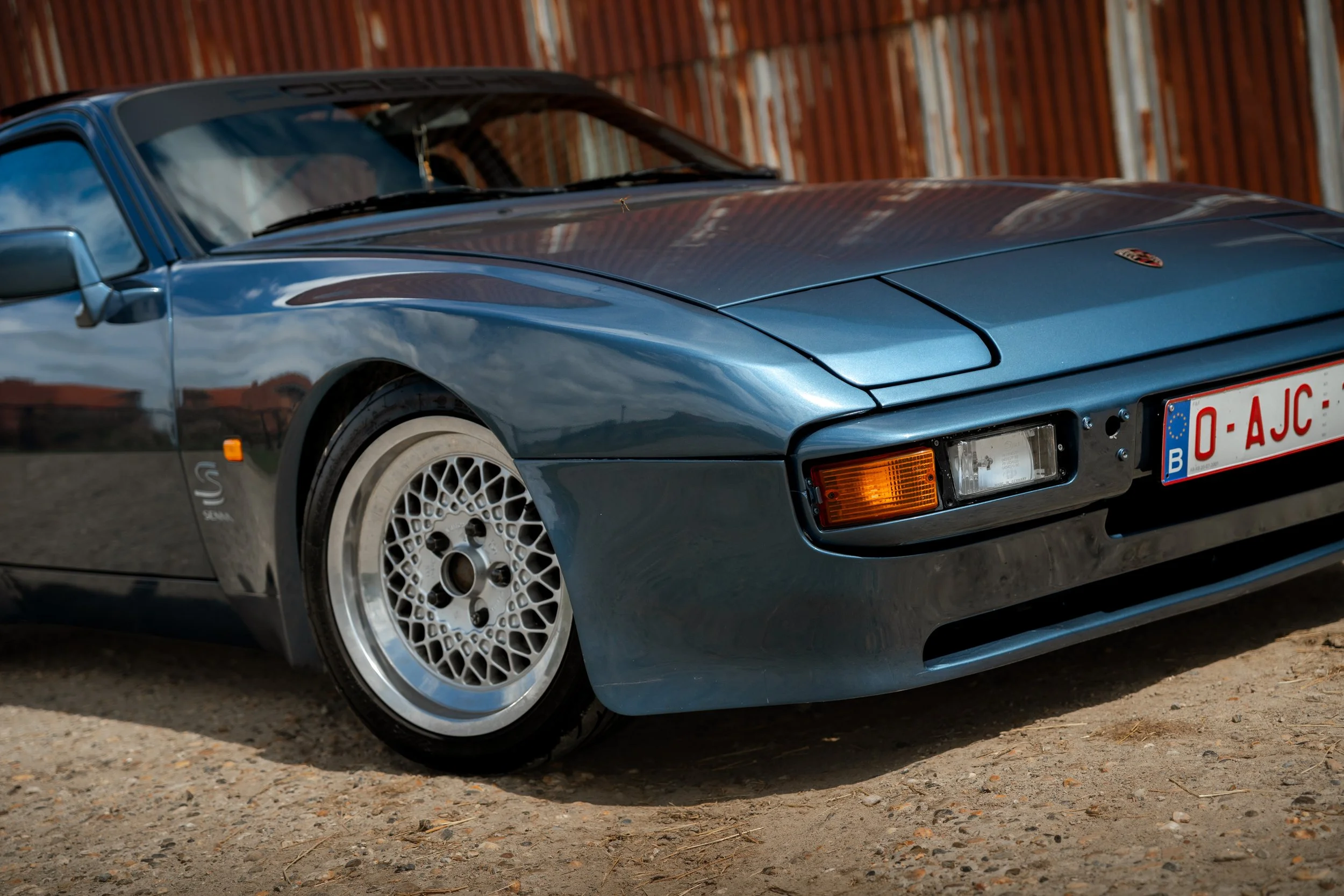 A blue vintage sports car with a European license plate parked on a gravel surface, with a wooden fence in the background.