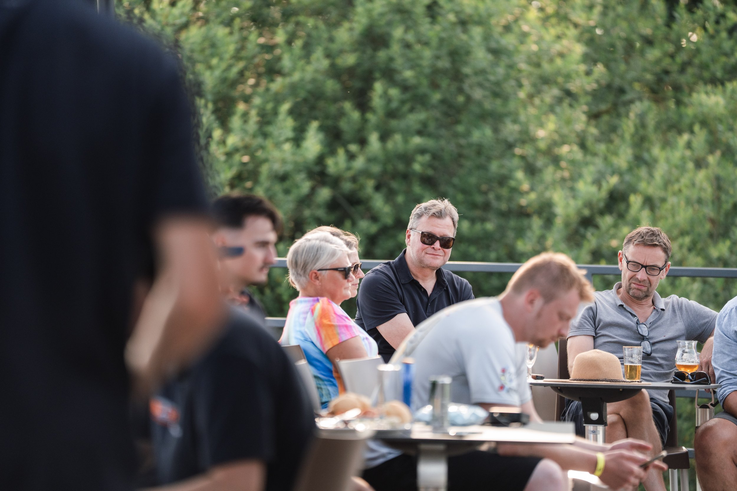A group of diverse people sitting outdoors at a social gathering, enjoying drinks and conversations on a sunny day with greenery in the background.