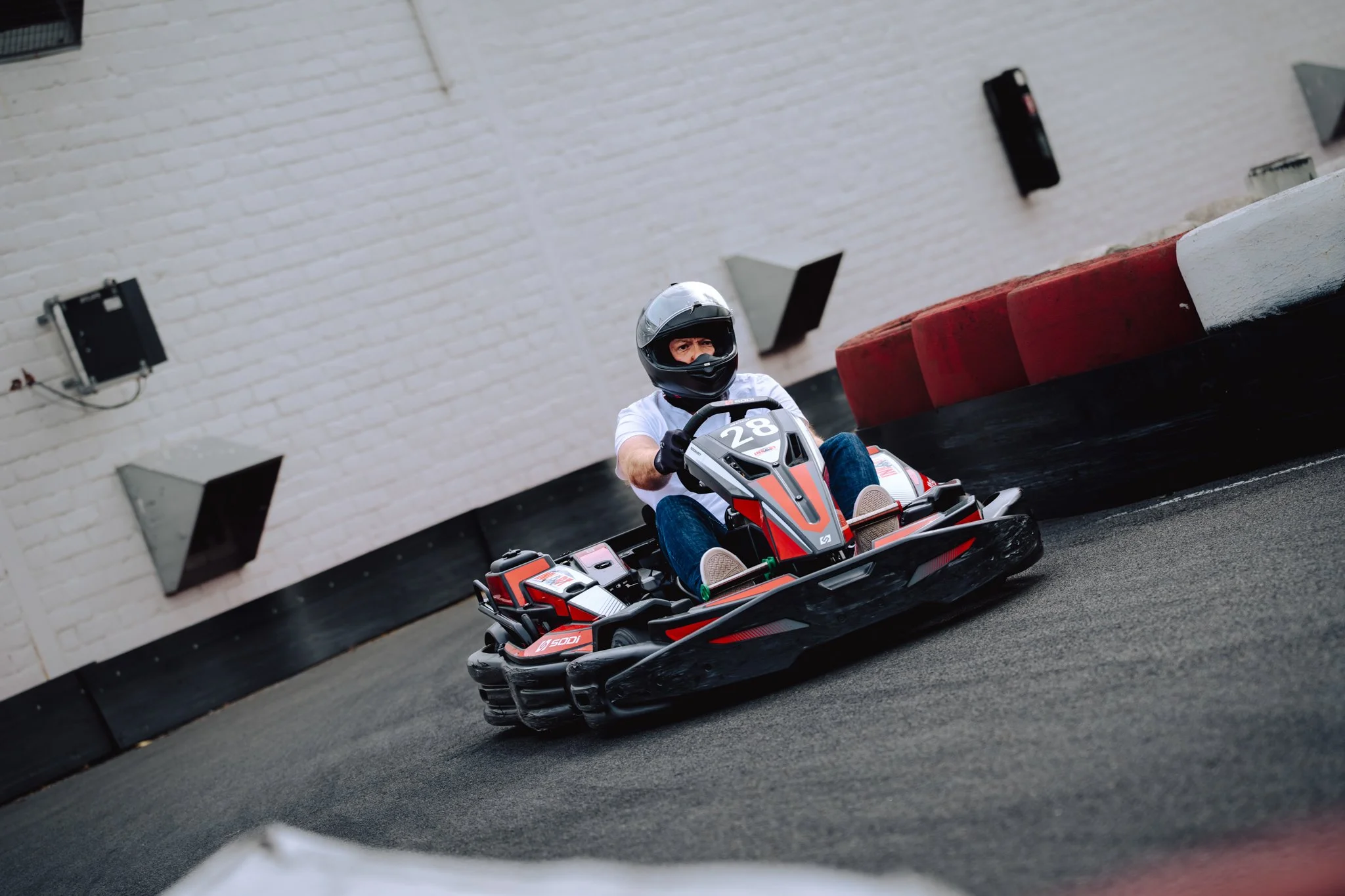 A person in a helmet driving a go-kart on an indoor track with a brick wall background and red and white barriers.