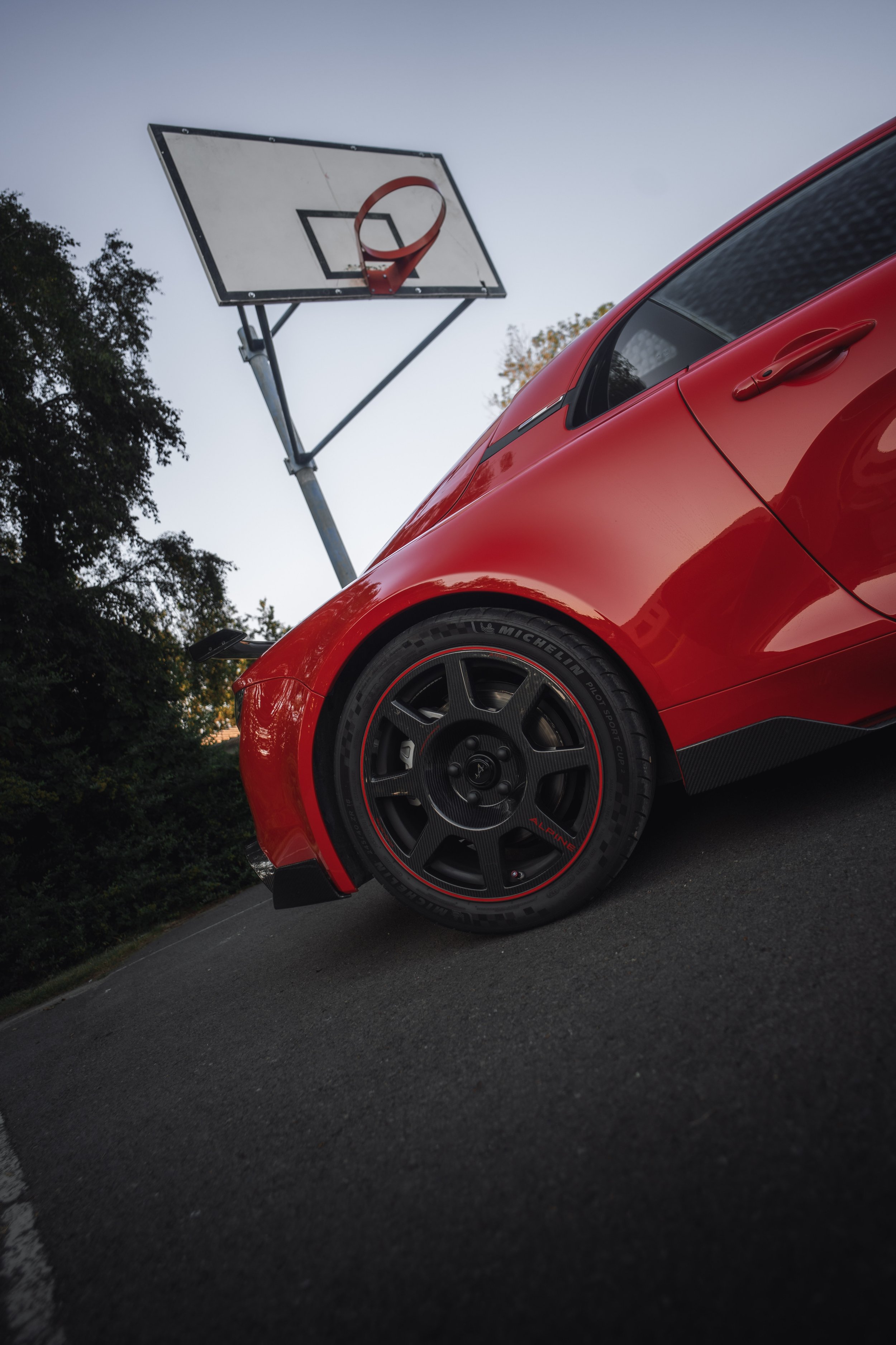 A red sports car parked on the side of the road with a basketball hoop in the background and trees on the side.