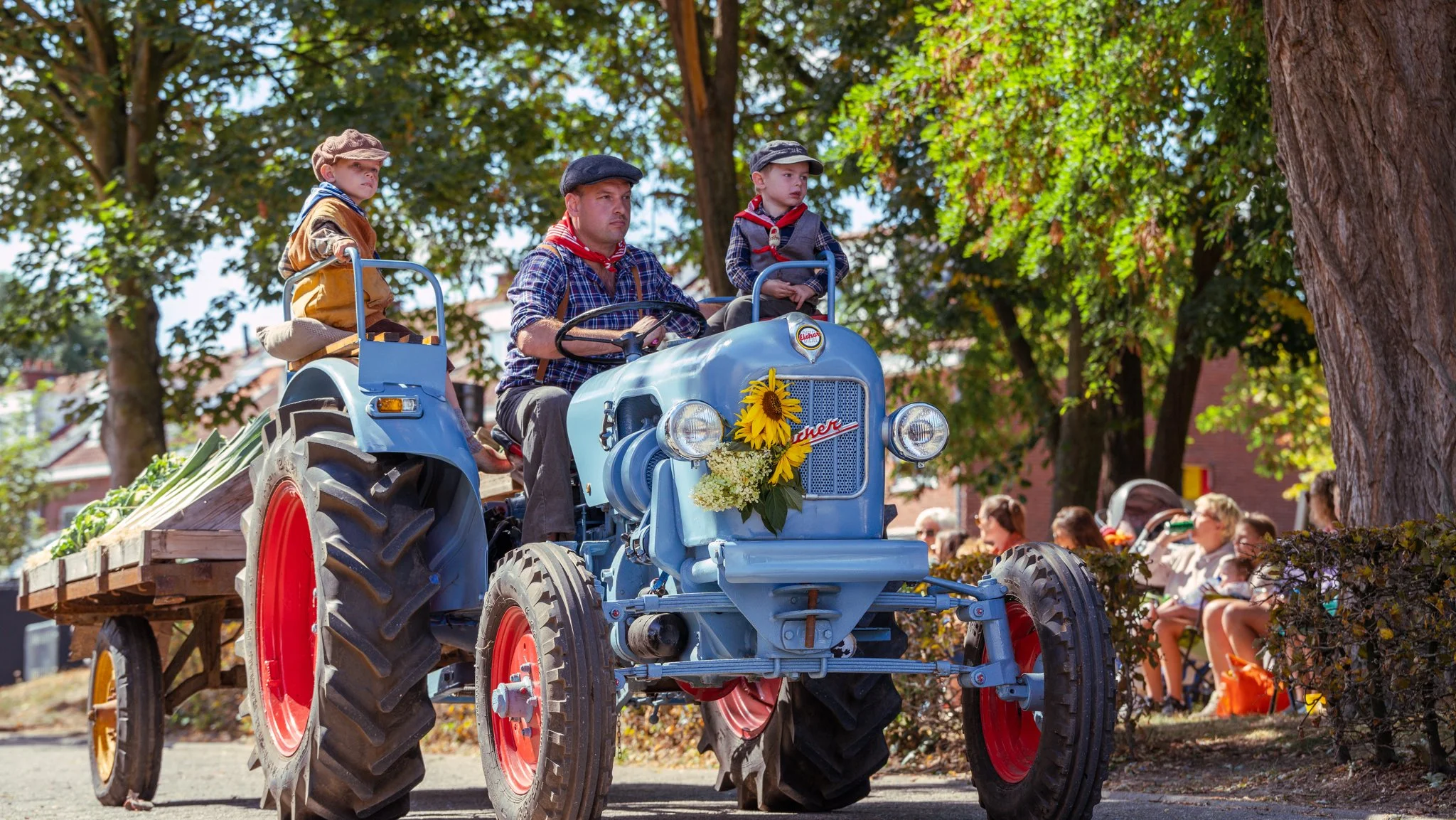 A man and two children riding a vintage blue tractor decorated with sunflowers, pulling a cart with vegetables during a parade or festival, with people sitting and watching in the background under trees.