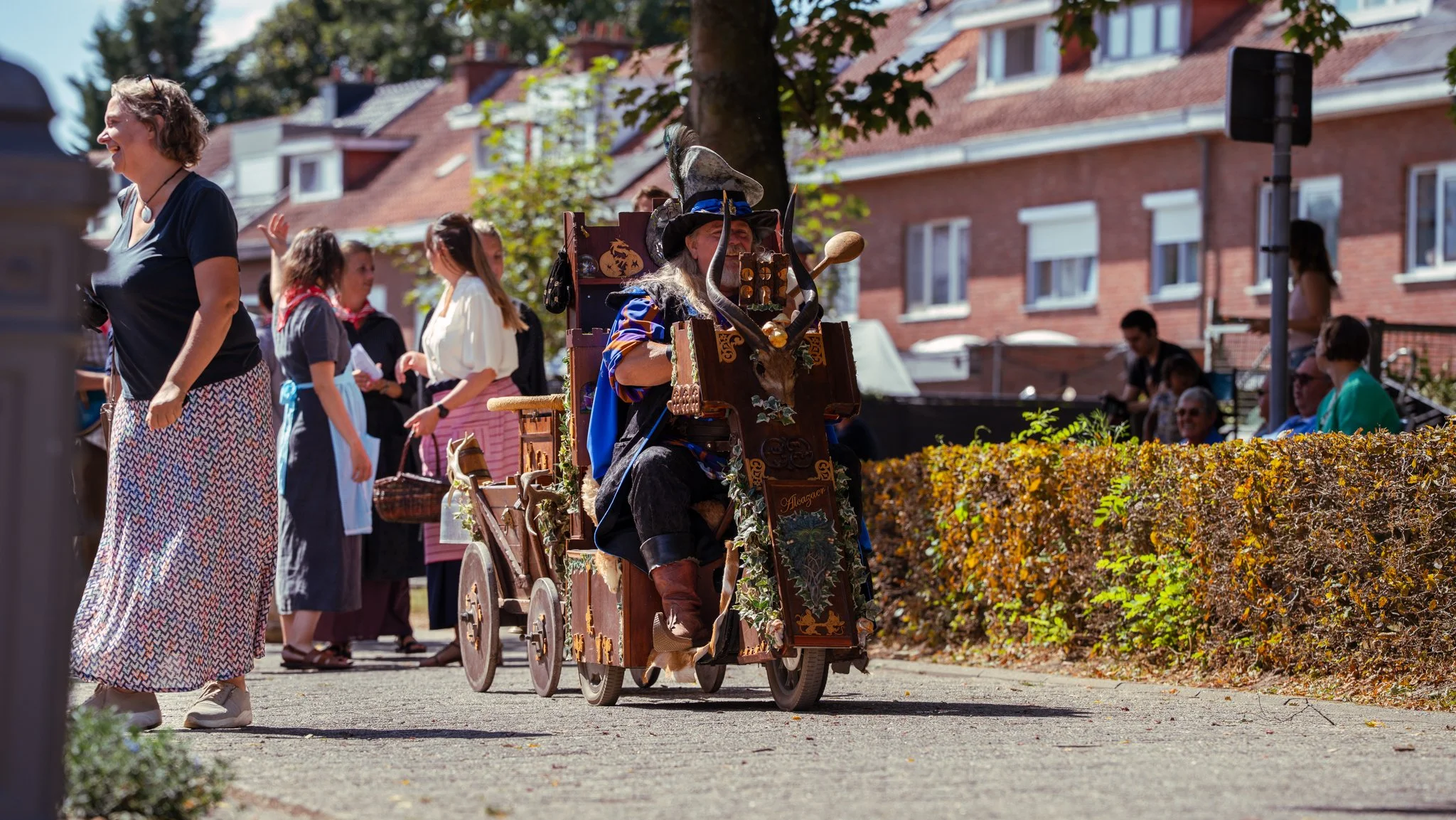 A person dressed as a wizard with a goat skull on their head riding a decorated wooden cart through a themed outdoor event, with people in vintage clothing watching and smiling in the background.