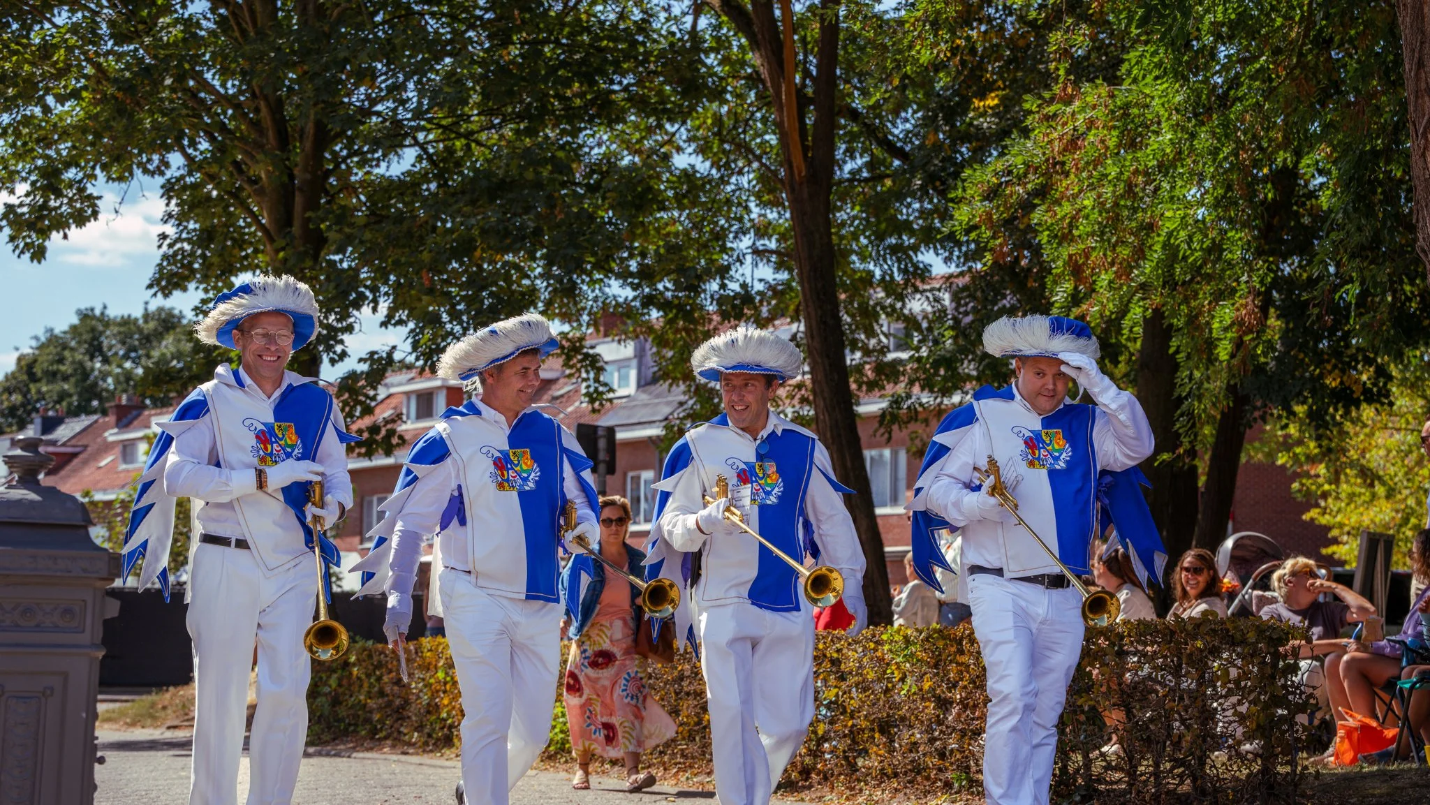 Four men dressed in elaborate costumes with white and blue clothing, feathered hats, and playing trumpets, walking outdoors in a parade or festival setting, with trees, spectators, and buildings in the background.