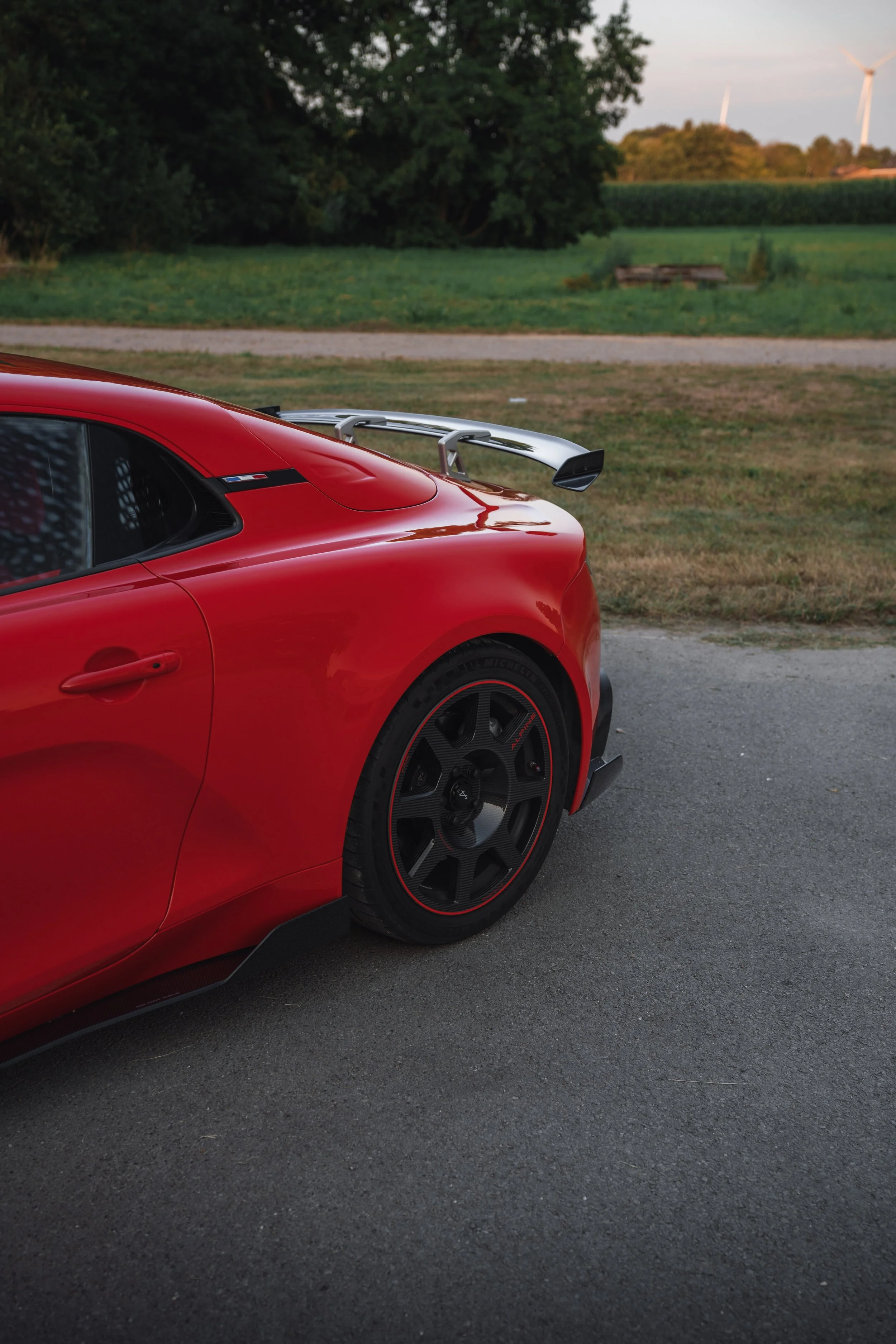 Close-up of the back part of a red sports car with a rear wing, parked on the side of a rural road with green fields and wind turbines in the background.