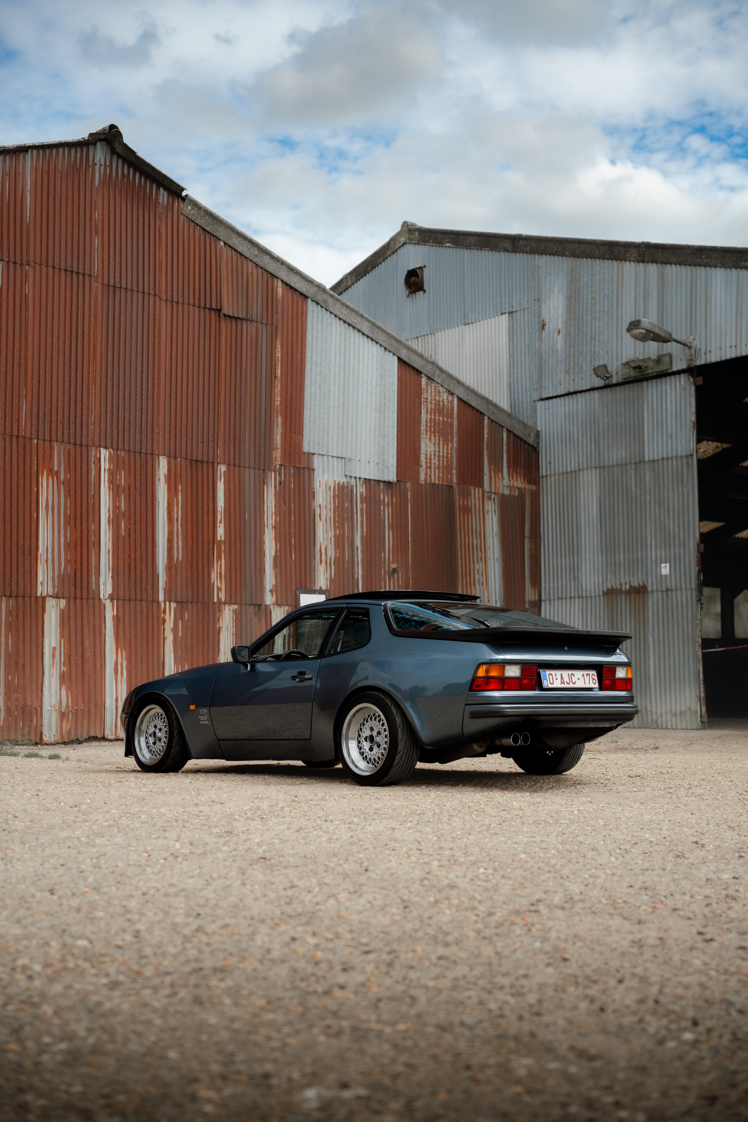 A dark gray Porsche 944 parked on a gravel lot in front of a weathered red and gray industrial building under a cloudy sky.