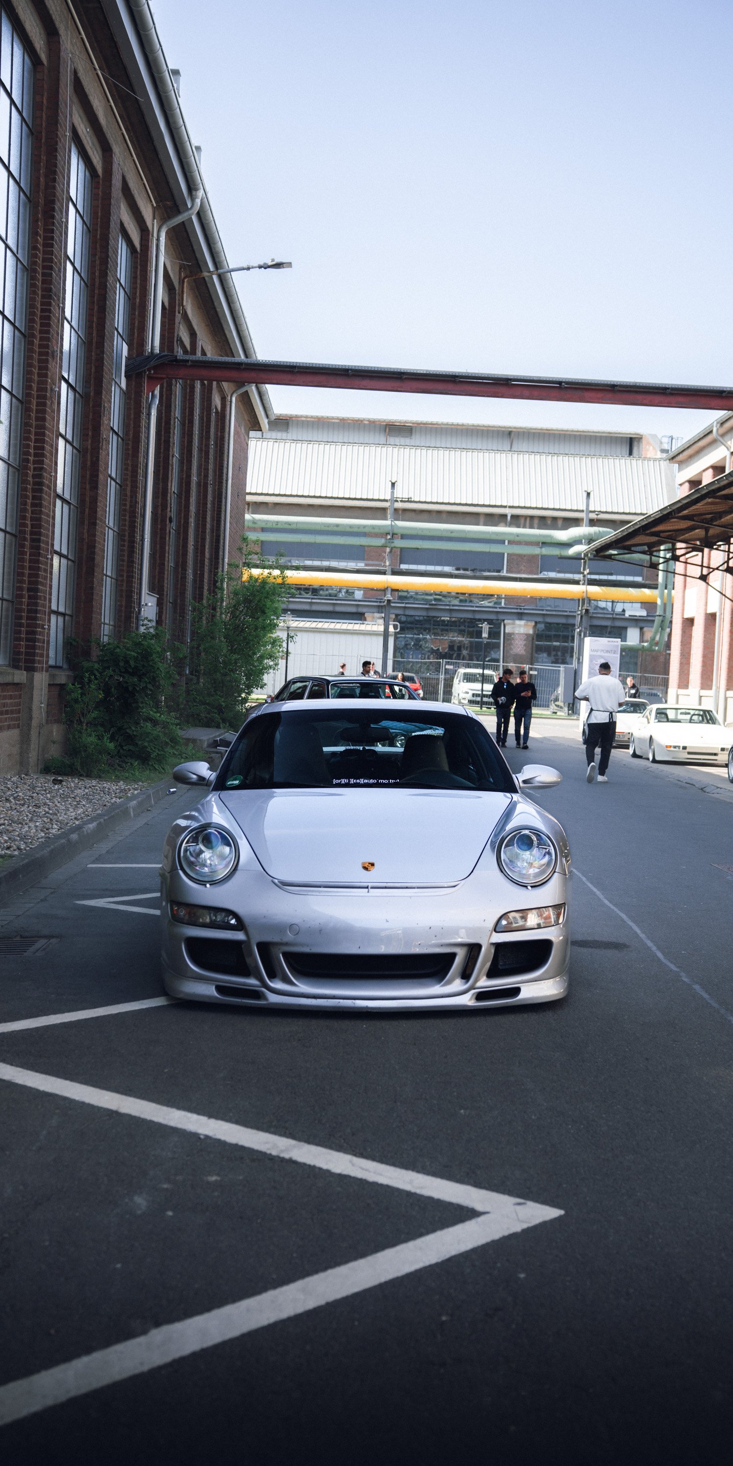 A silver Porsche sports car parked in a parking lot next to a brick building, with people and vehicles visible in the background.