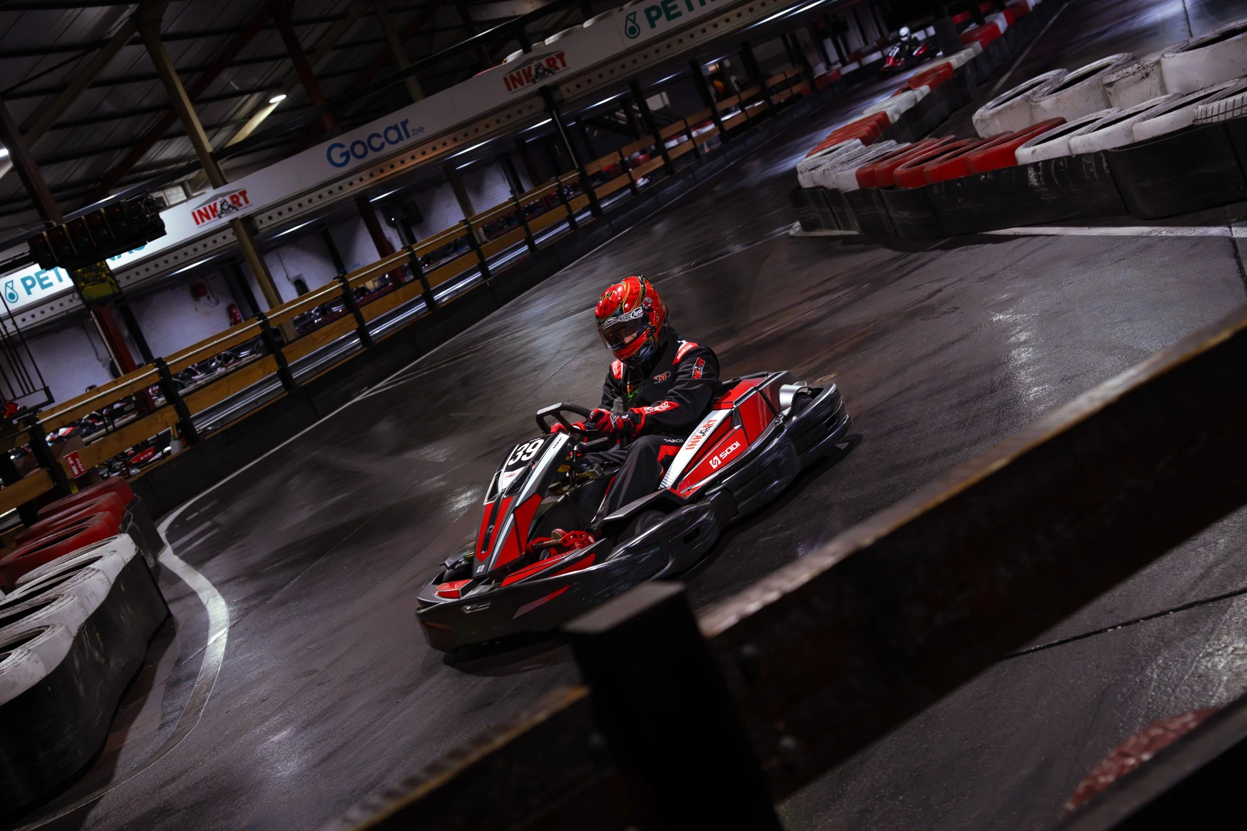 A person driving a go-kart on an indoor race track, wearing a red helmet and black racing suit, with a wet pavement and orange safety barriers around the track.