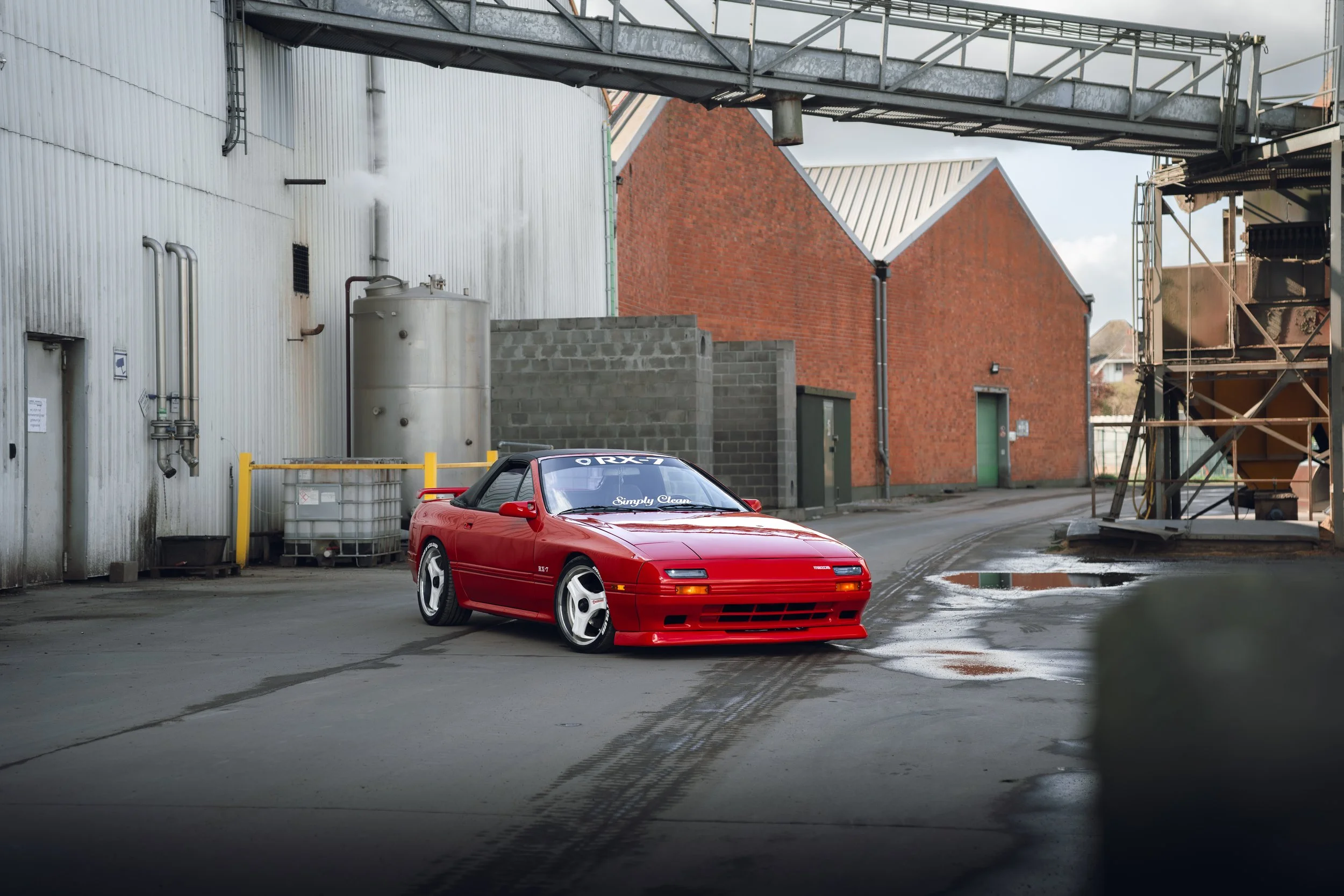 Red sports car with black roof parked in an industrial alleyway with red brick buildings, a corrugated metal building, and puddles on the ground.