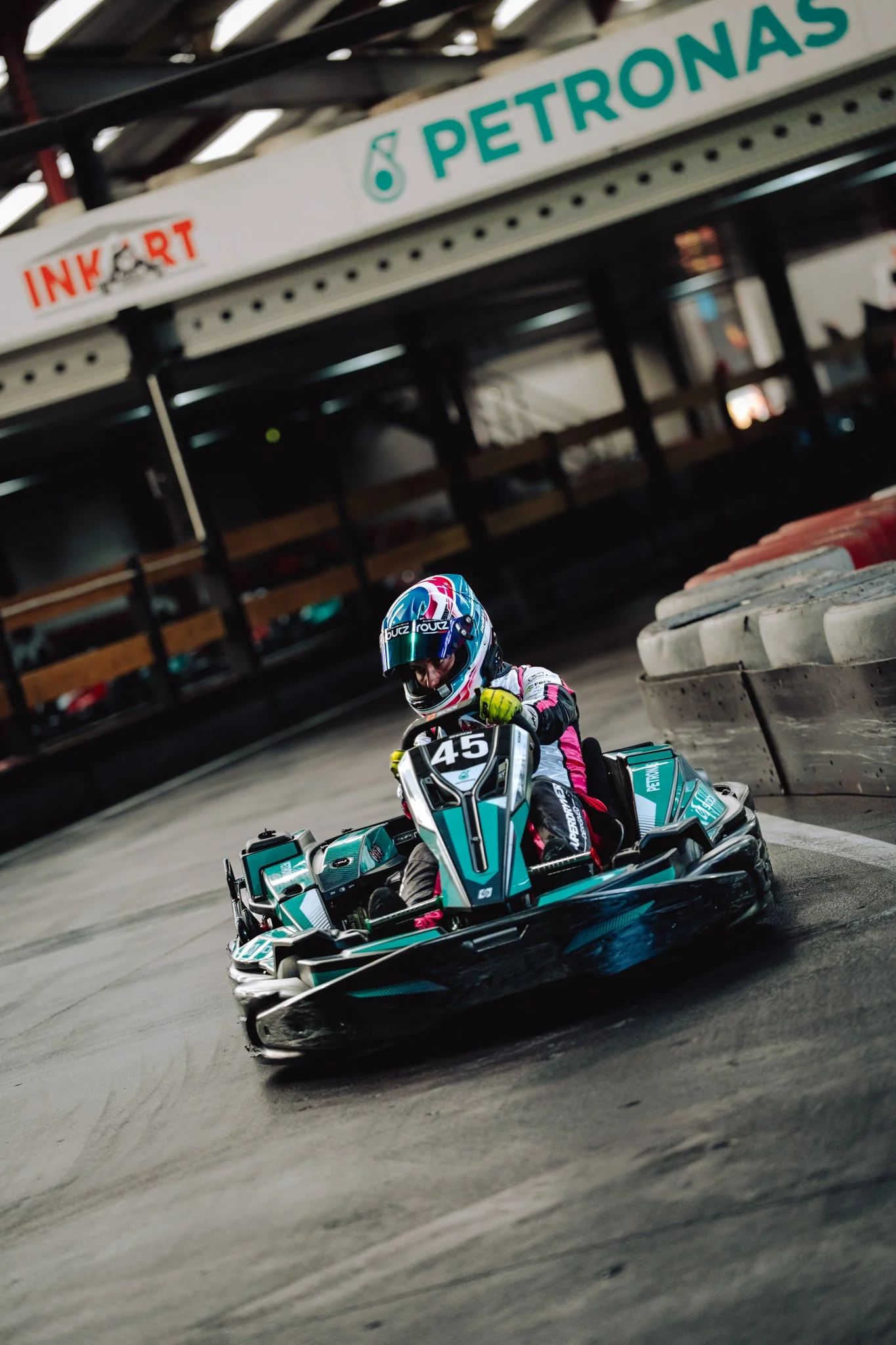 A person racing in a go-kart on an indoor track, wearing a helmet and racing suit, with the number 45 on the front of the kart, and signs for PETRONAS and INKART visible above the track.