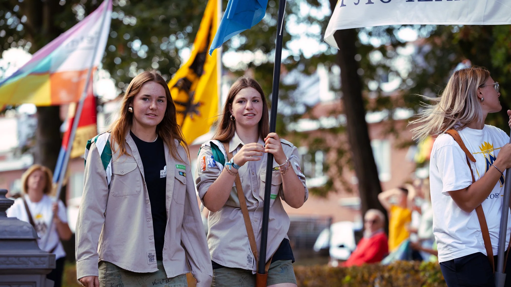 Three young women, dressed in Girl Scout uniforms, are participating in a parade or outdoor event, holding flags and walking among a crowd. The background features trees and out-of-focus people.